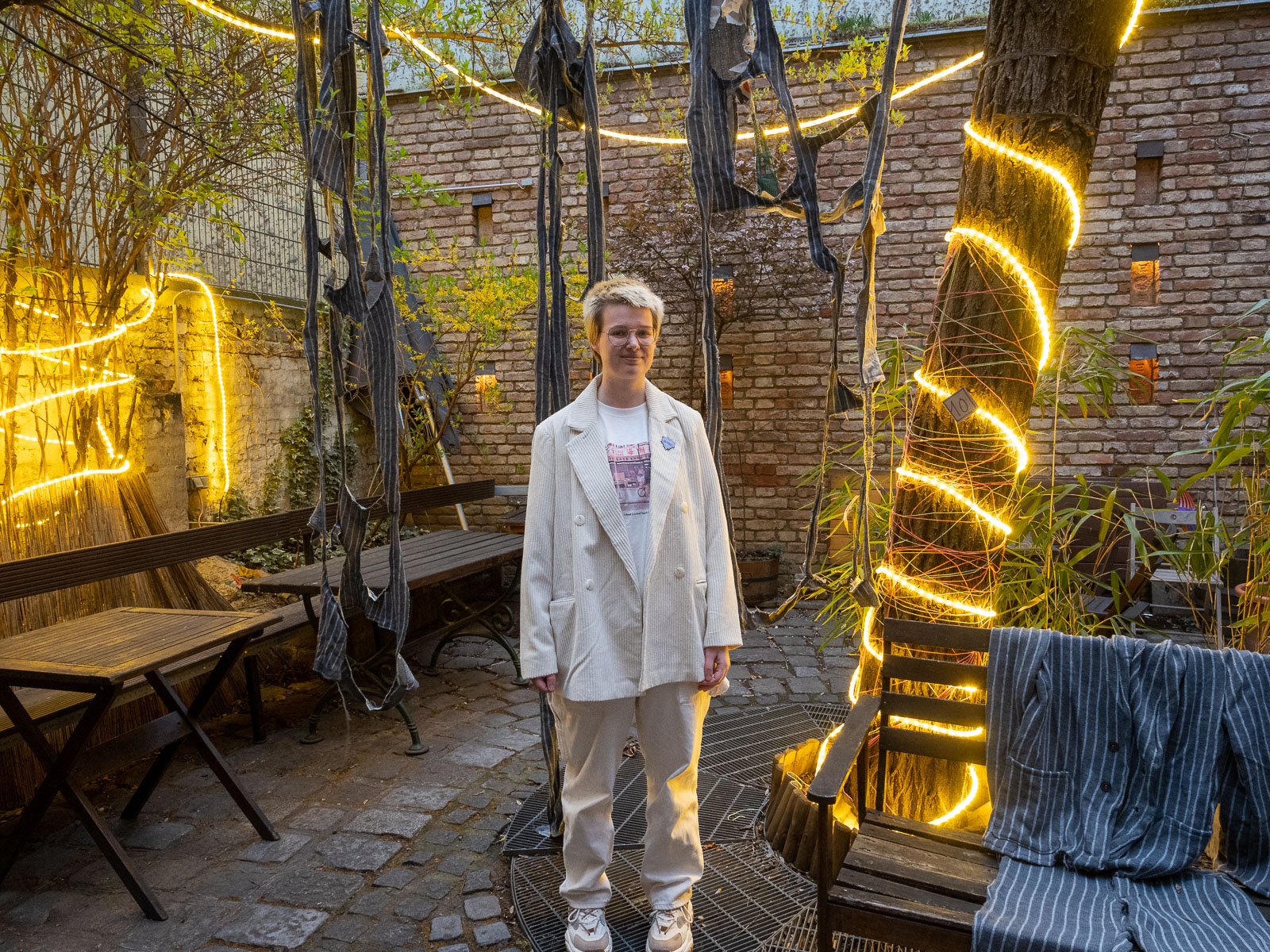 A young transgender man in a white corduroy suit in front of his artwork, a knitted grey and white striped suit, next to a tree wrapped in lights