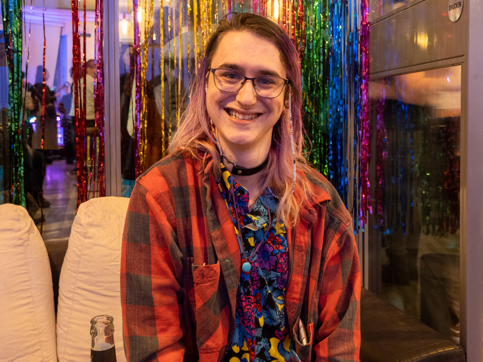 Portrait of a transgender/nonbinary artist (she/they) in front of a glitter curtain, dressed in a red and black checkered shirt. 