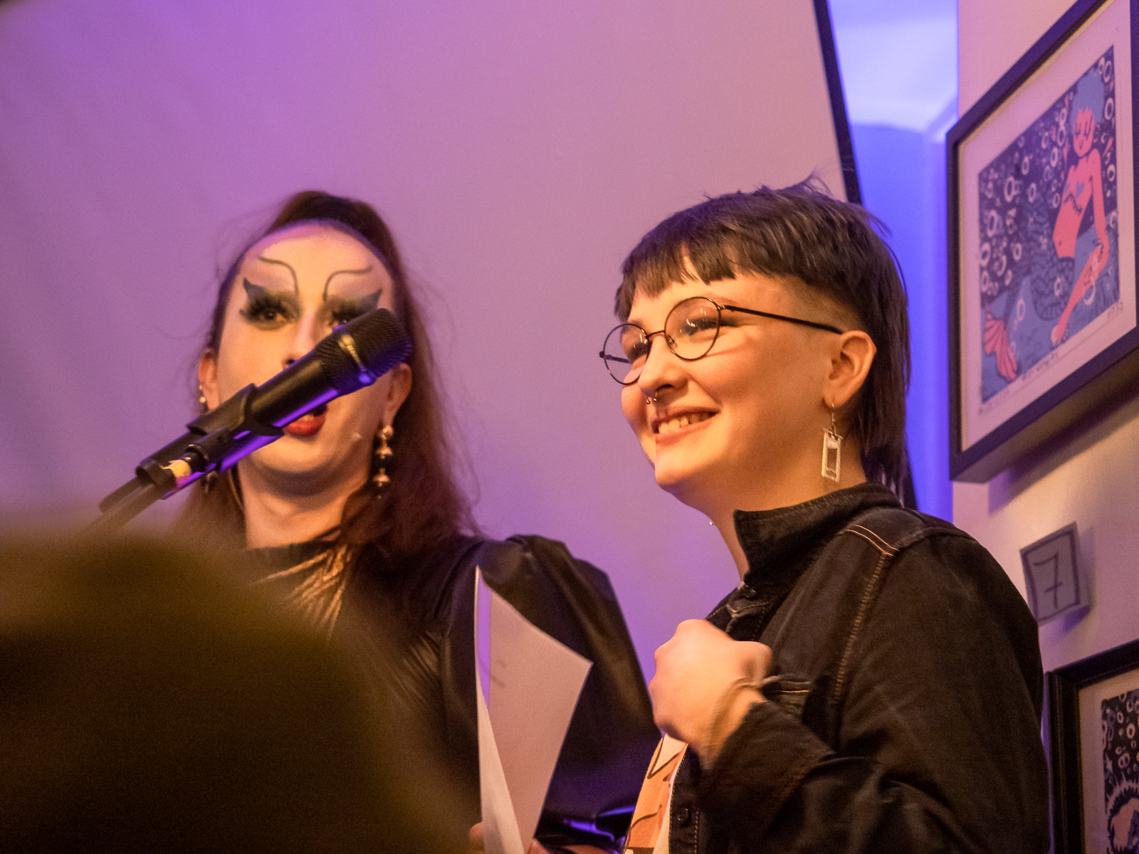 two artists on stage, one with makeup, the other with glasses, smiling