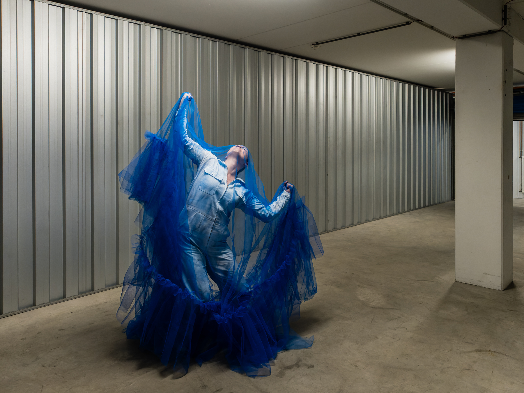 Actress in a white overall covered in a bright blue full body transparent veil, dancing in an empty storage room.