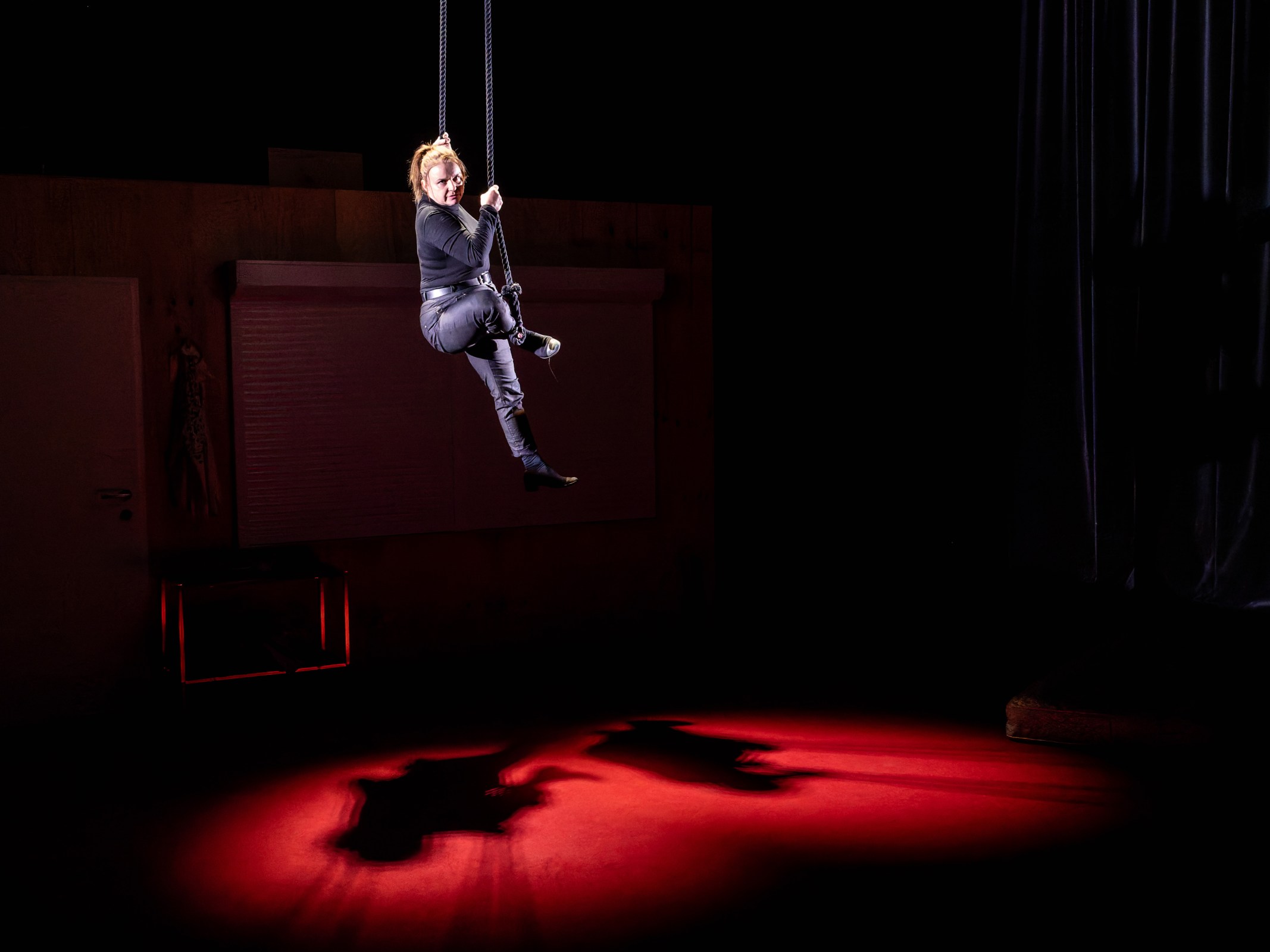A performer gracefully suspended from ropes, dressed in black, in a dimly lit space with a red spotlight illuminating the floor and casting a shadow.