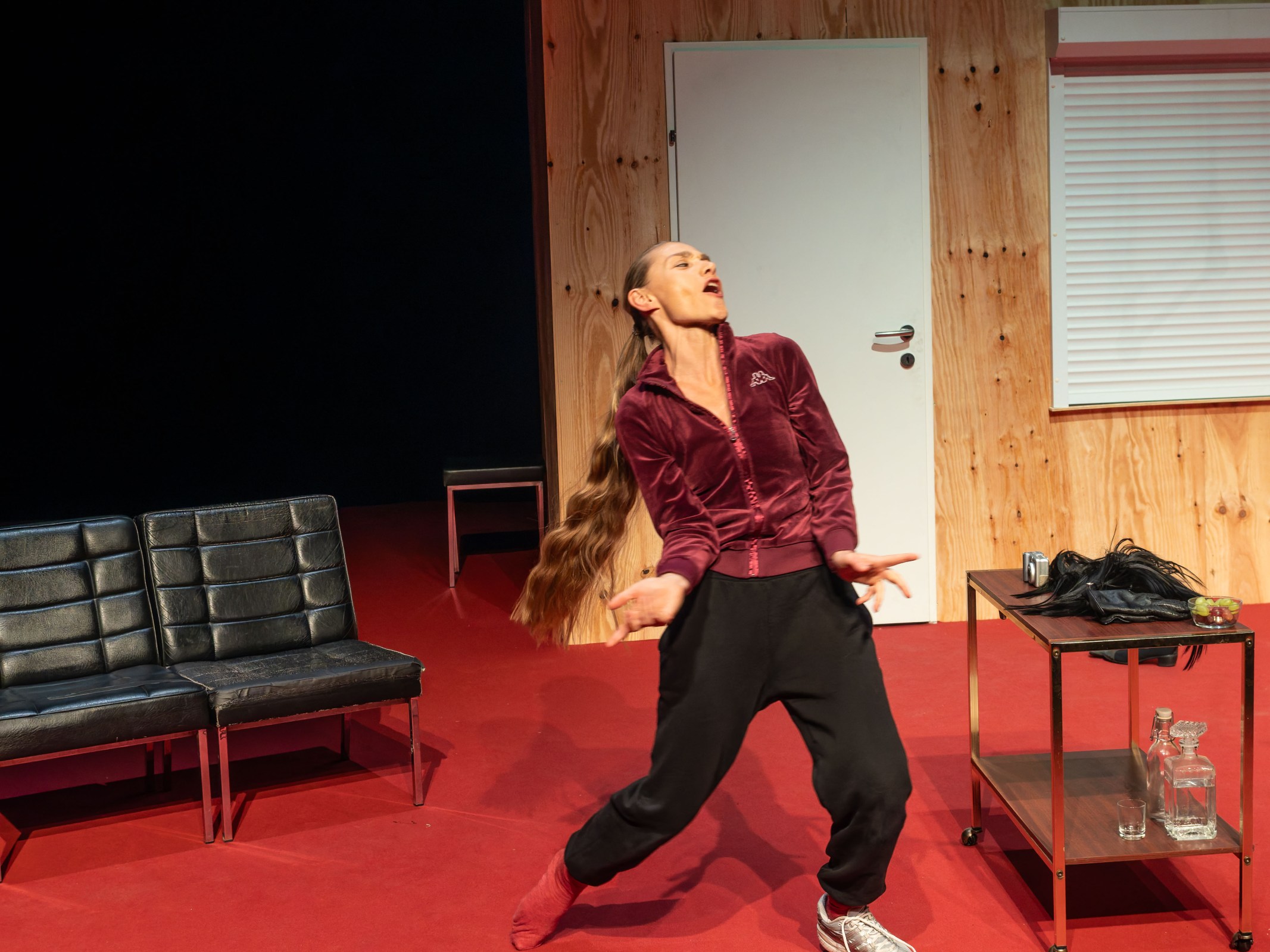 A performer in a burgundy tracksuit and black trousers expresses emotion on stage, surrounded by minimalist furniture, including two black chairs and a small table with a bottle, glass, and grapes.