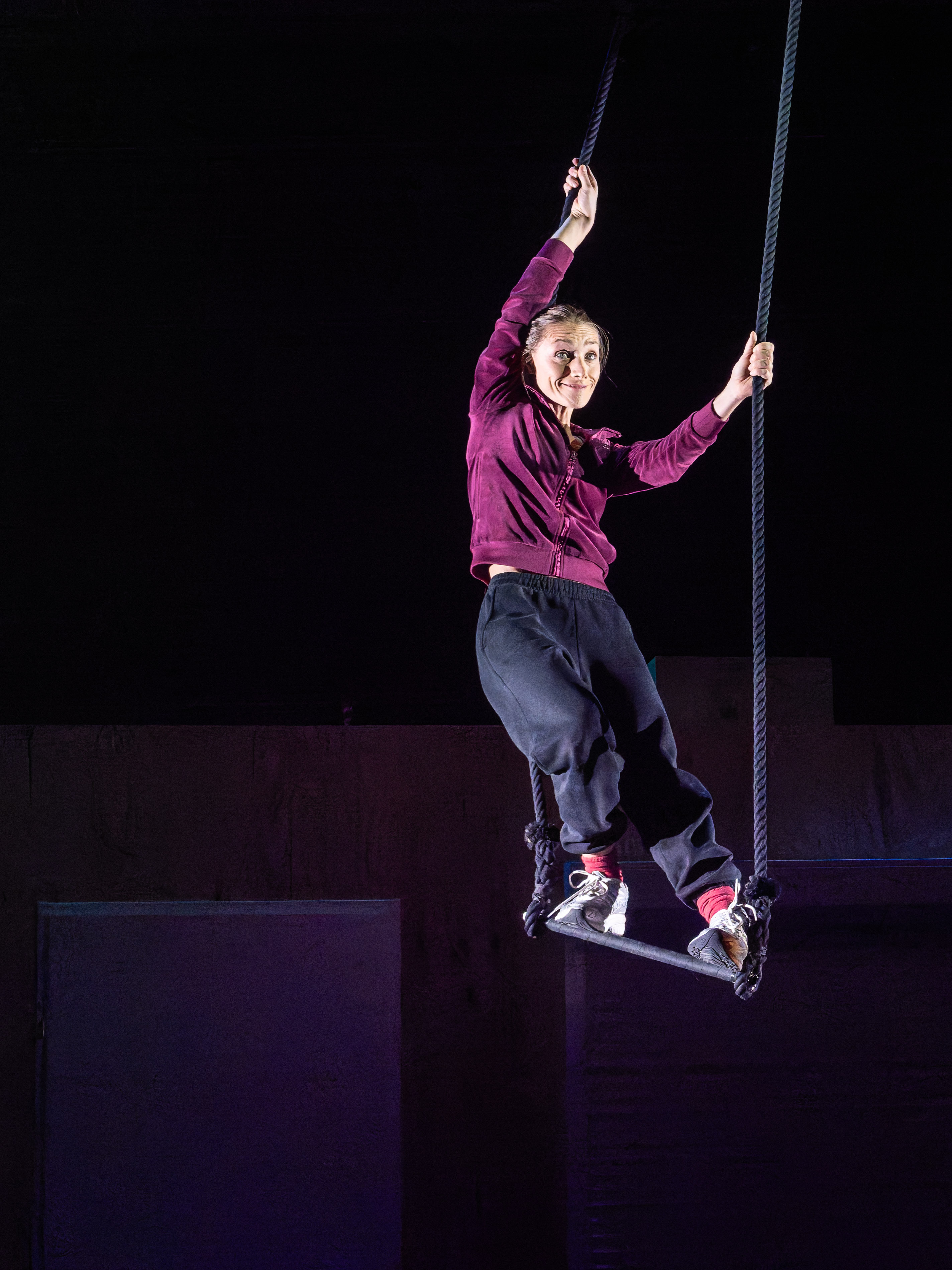 A performer hanging from a rope on a swing, dressed in a purple top and black trousers, with a playful expression against a dark background.