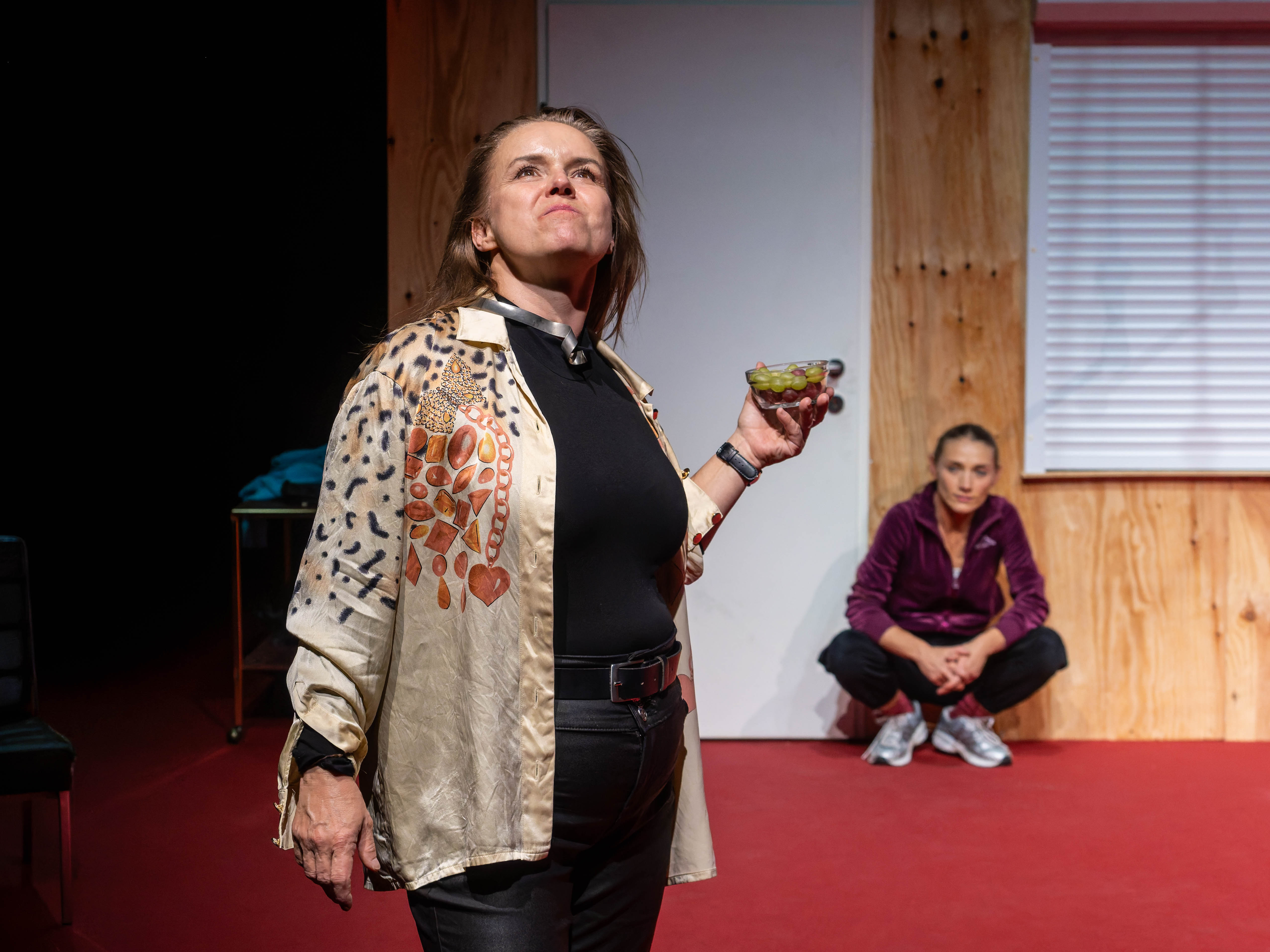 A woman in an elaborately patterned shirt holds a bowl of grapes and looks defiantly upward, while another woman in a purple tracksuit sits in the background, observing attentively. The setting features a wooden wall and red flooring, creating a dramatic atmosphere.