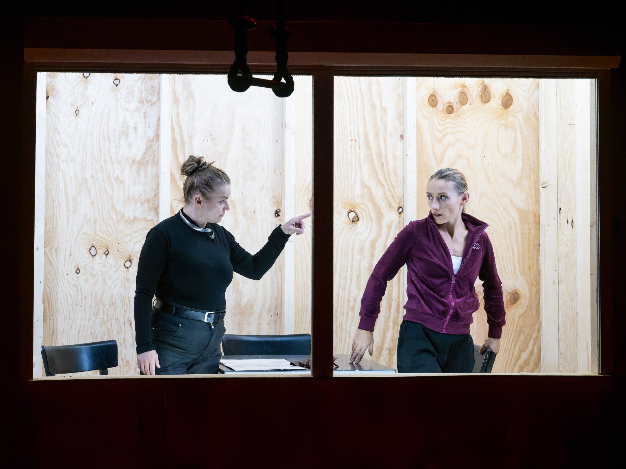 Two women engaged in an intense conversation inside a wooden-framed room, one is pointing while the other is standing with a tense posture.