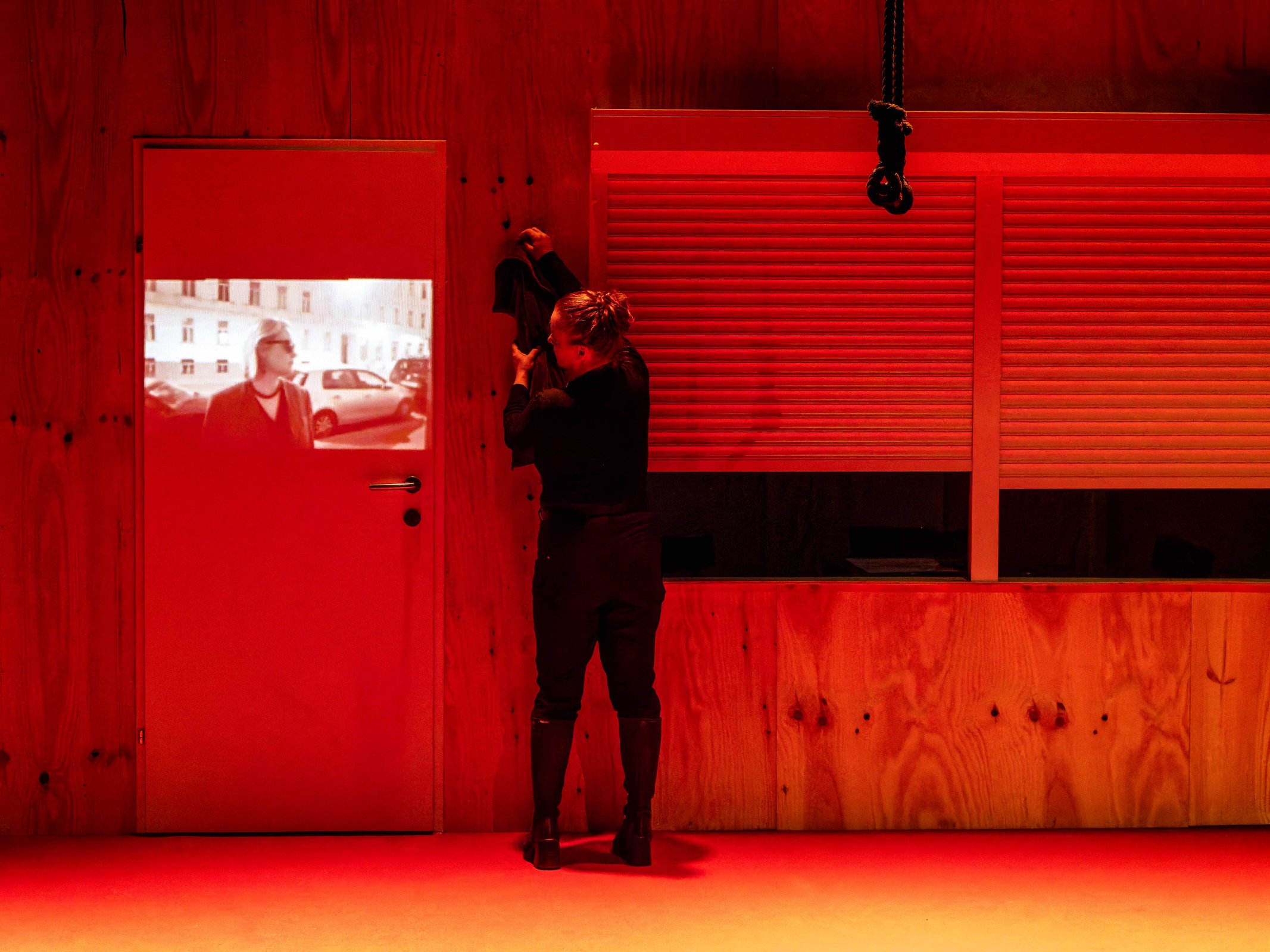 A woman wearing black clothing is interacting with a dark door featuring a projected image of a man in a city setting. The scene is illuminated with red lighting, creating a dramatic atmosphere.