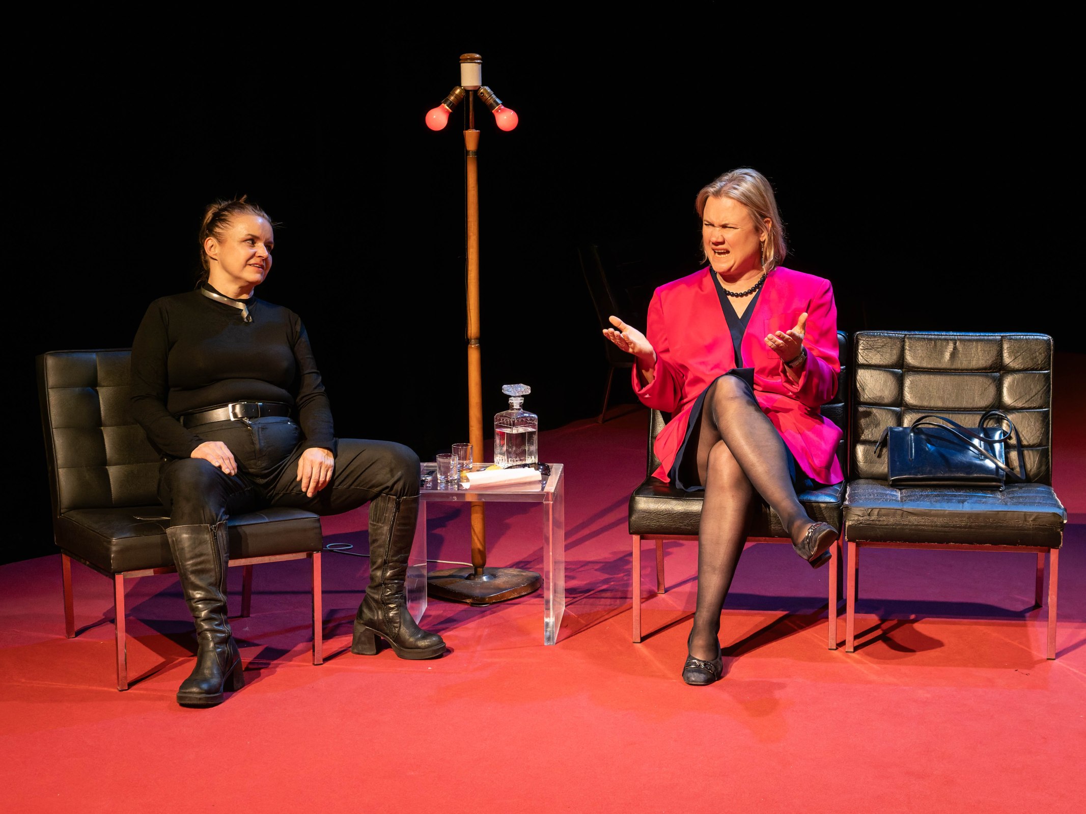 Two women seated on black chairs in a stage setting, engaged in conversation. One woman is wearing a black outfit with knee-high boots, while the other is in a bright pink blazer and black dress. A side table holds a glass and a decanter, and a lamp with red bulbs stands behind them.