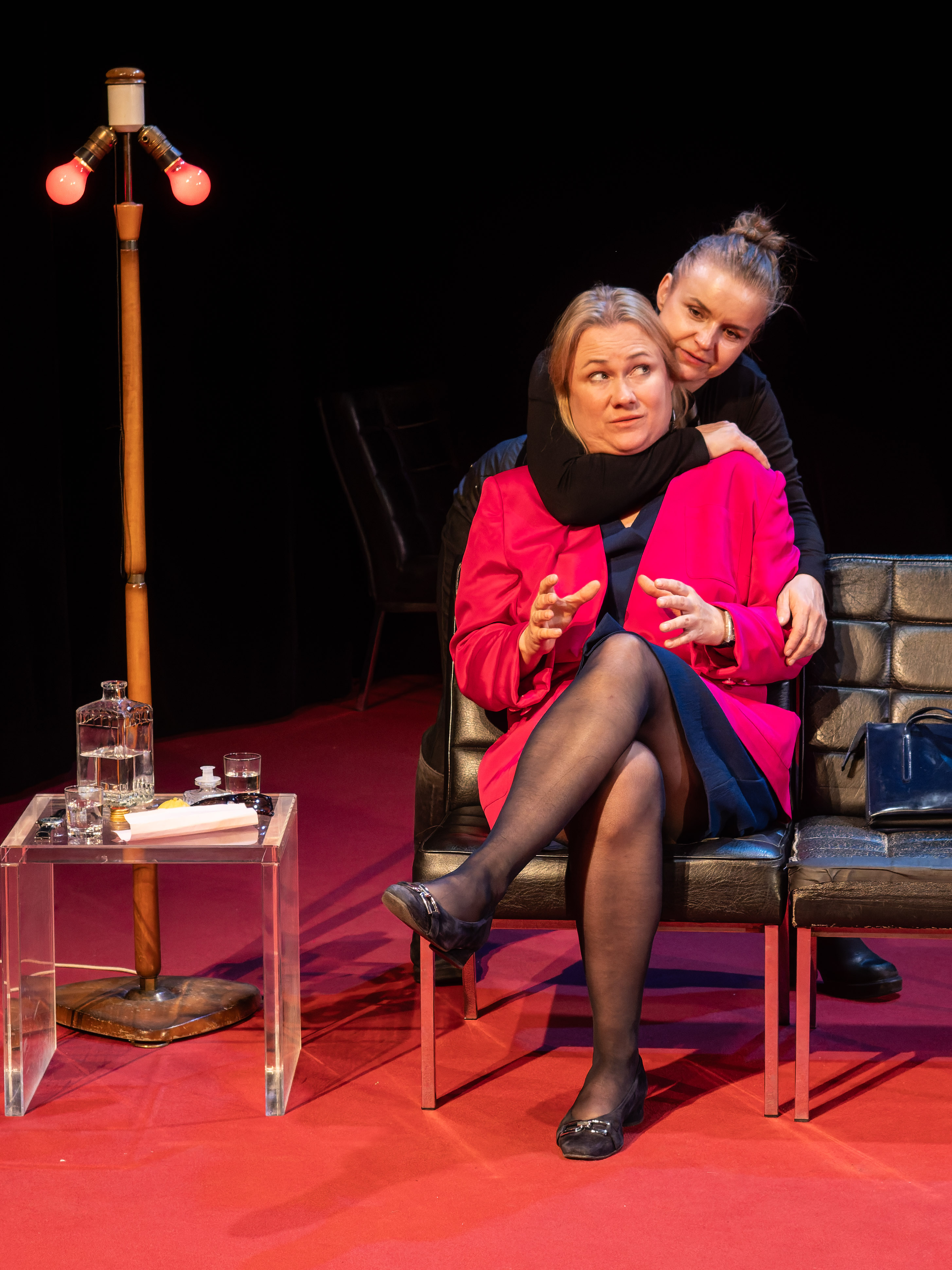 Two women seated on black leather chairs in a theatrical setting, with one leaning affectionately on the other. The woman in a pink jacket appears to be speaking, while the other woman, dressed in black, embraces her from behind. A stylish floor lamp with red bulbs and a small glass table with a decanter and glass sit nearby.