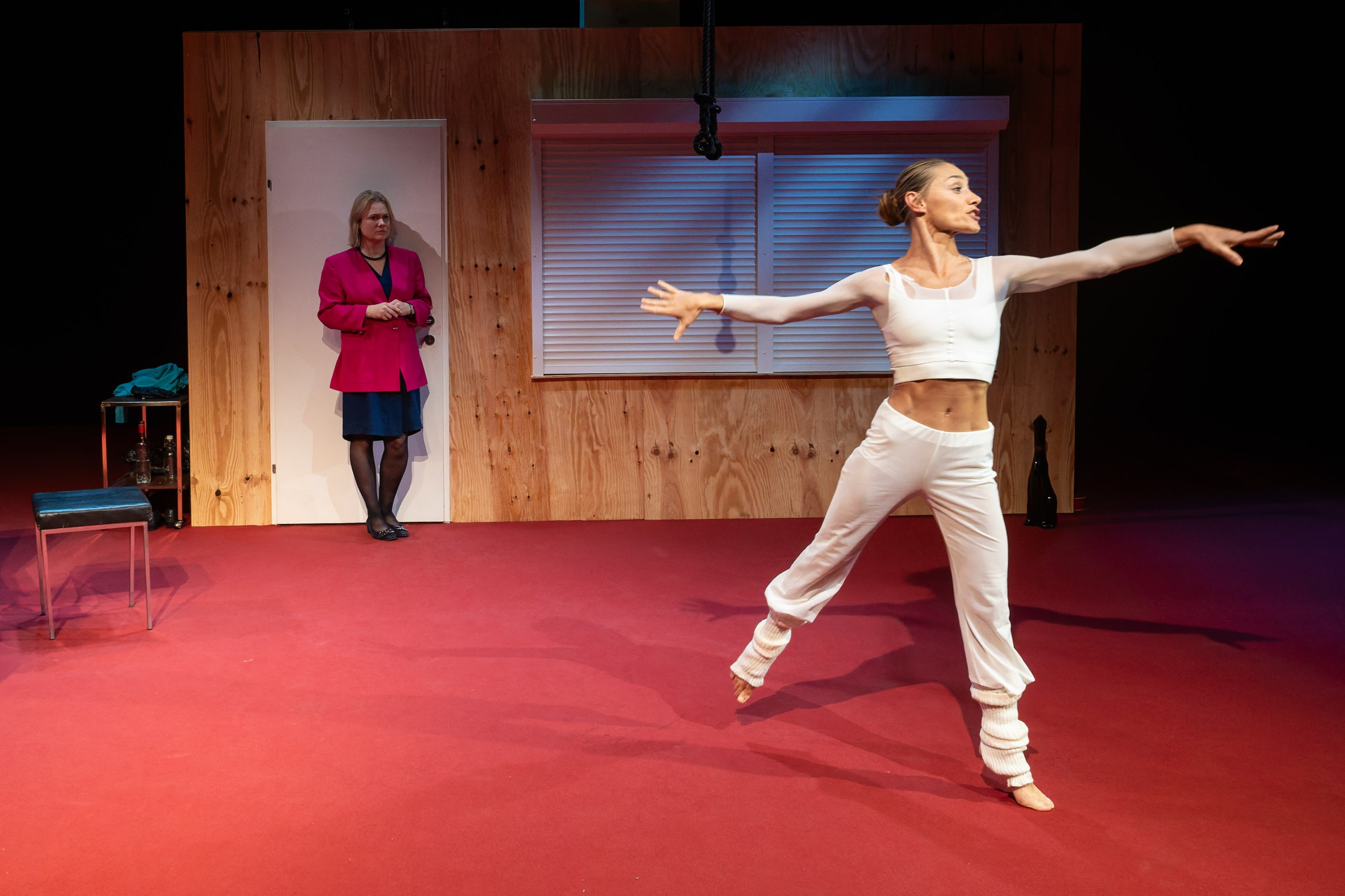 A dancer in a white outfit performs gracefully on stage, while a woman in a pink blazer looks on from the side.