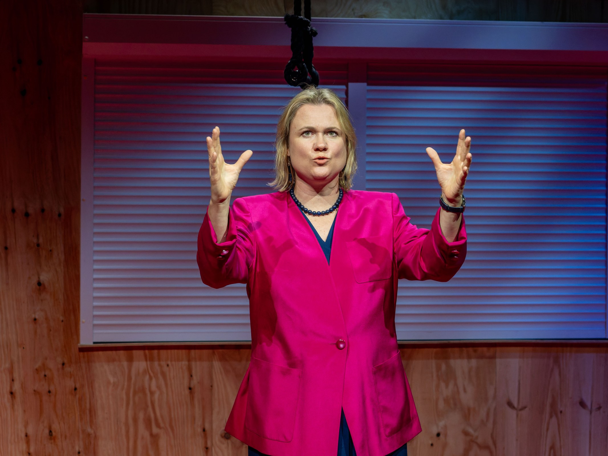 A woman in a bright pink blazer gestures expressively while speaking on stage, with a wooden backdrop and soft blue lighting.