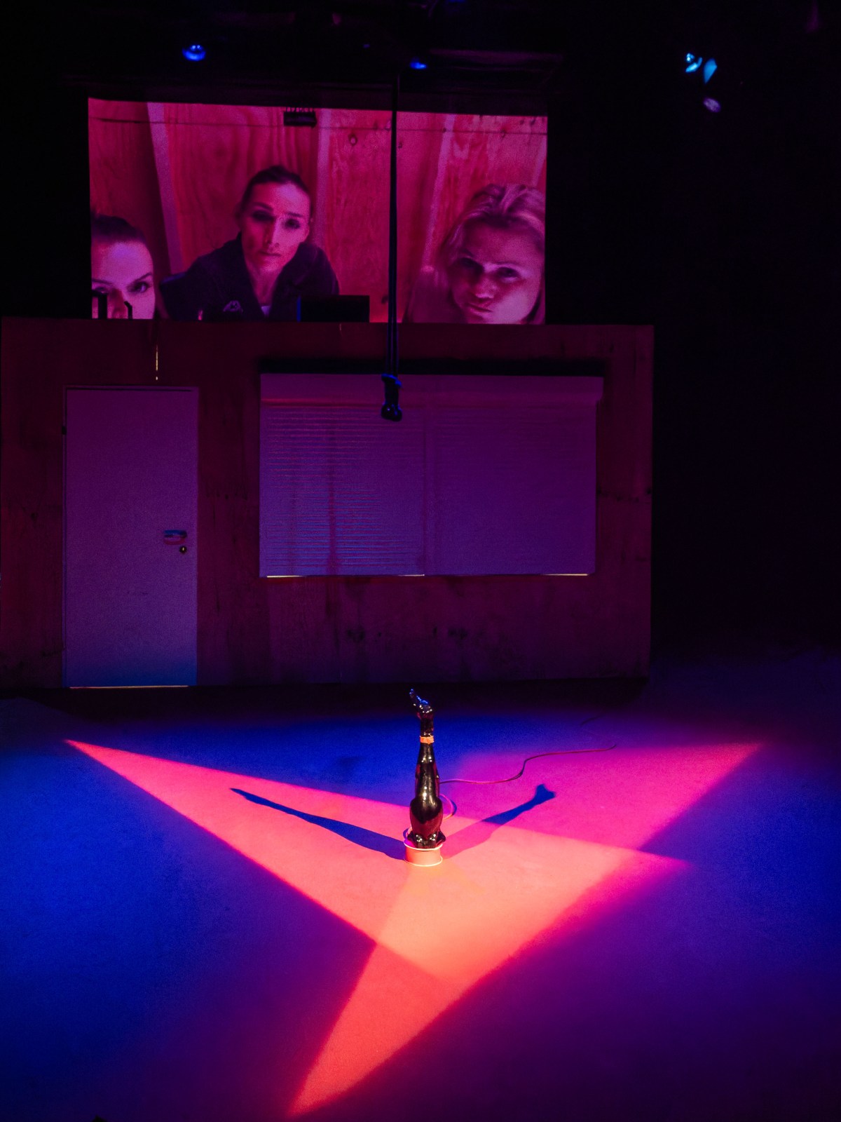 A dramatic stage scene featuring a spotlight on a champagne bottle with a hand sculpture next to it, set against a backdrop displaying faces of women in vibrant pink lighting.