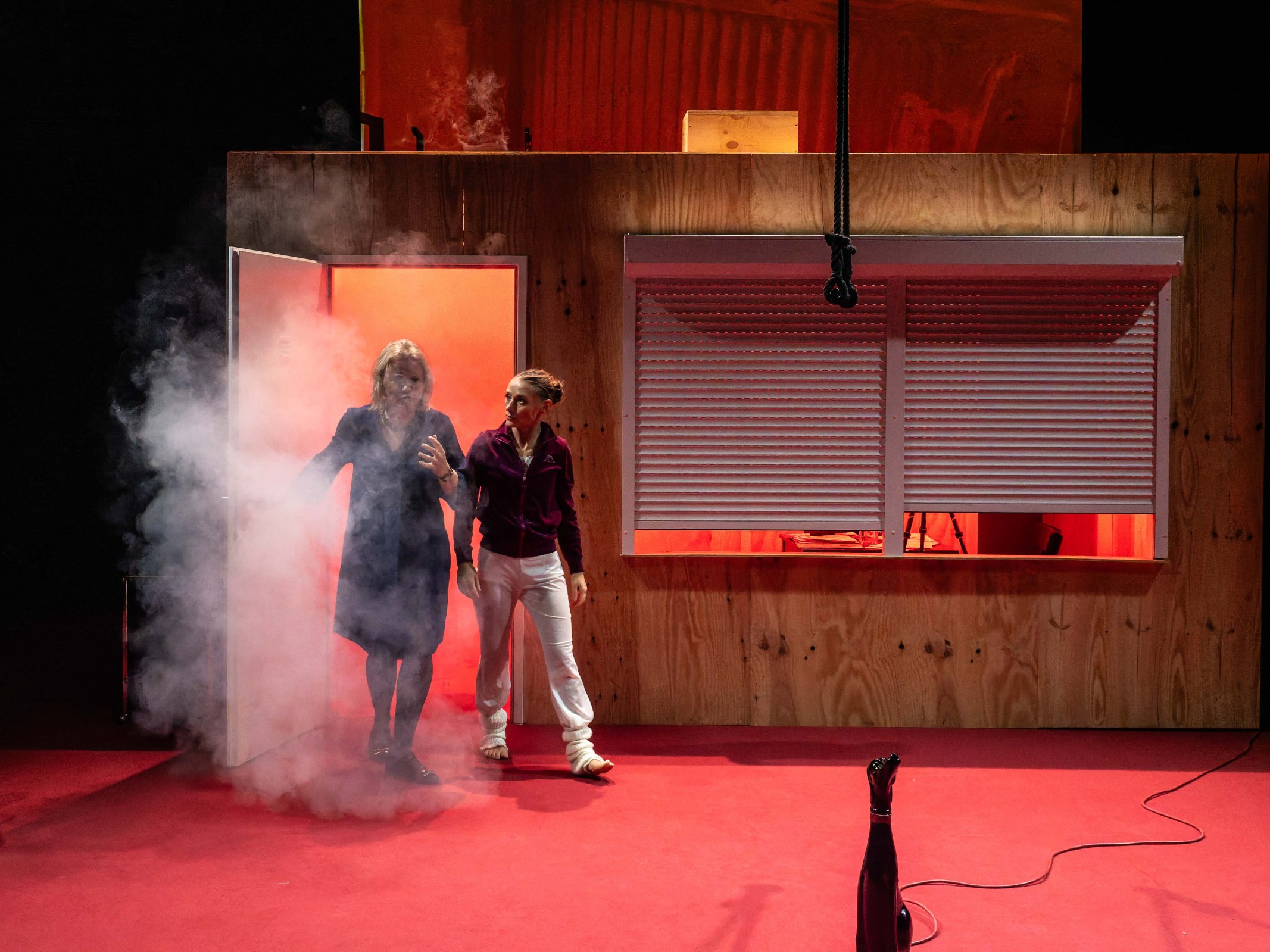 Two women emerging from a doorway, surrounded by smoke, on a stage set with a wooden house backdrop and a red carpet.