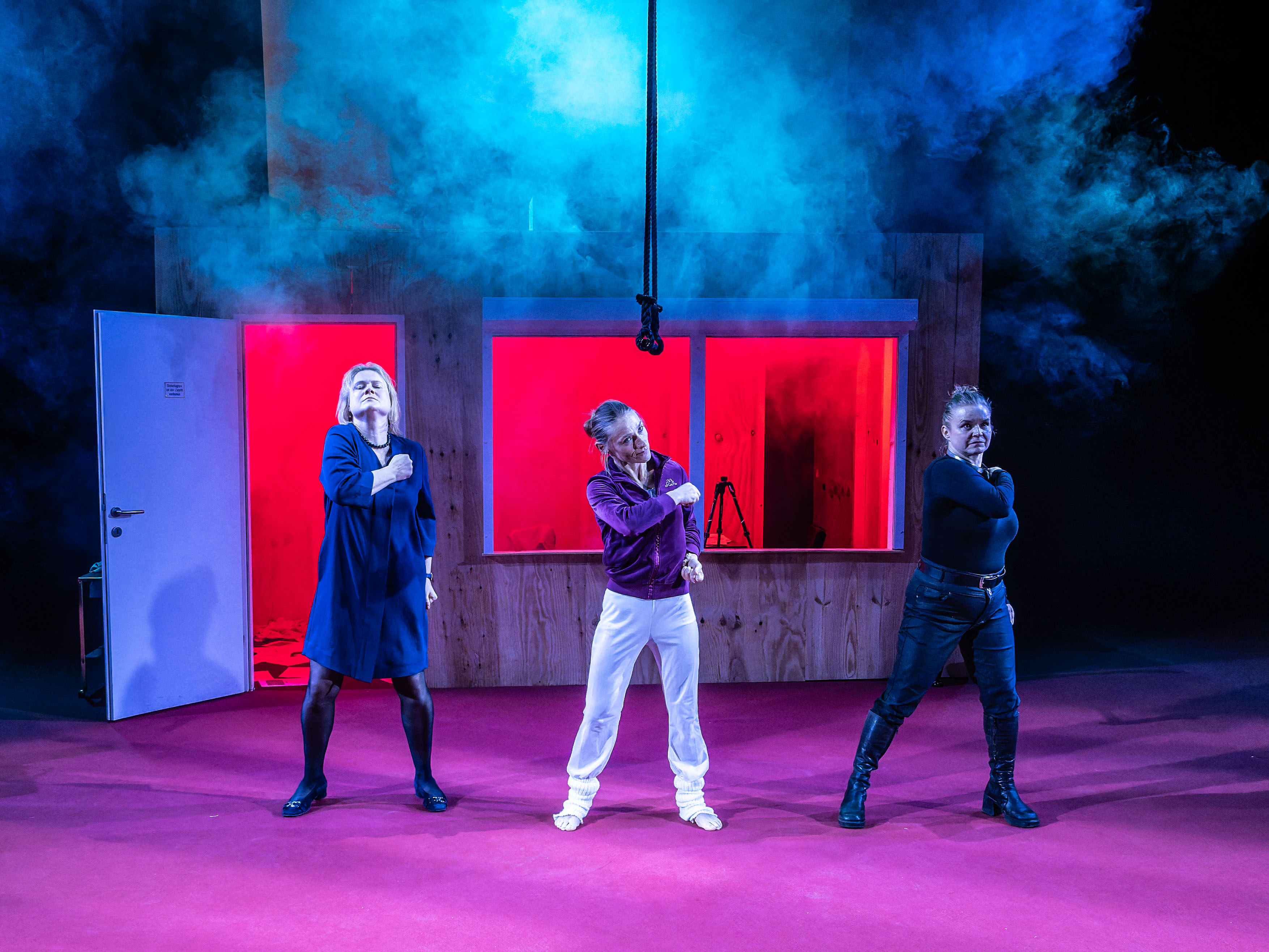 Three performers on stage striking a dramatic pose with a smoky background, featuring red-lit windows and a wooden set.