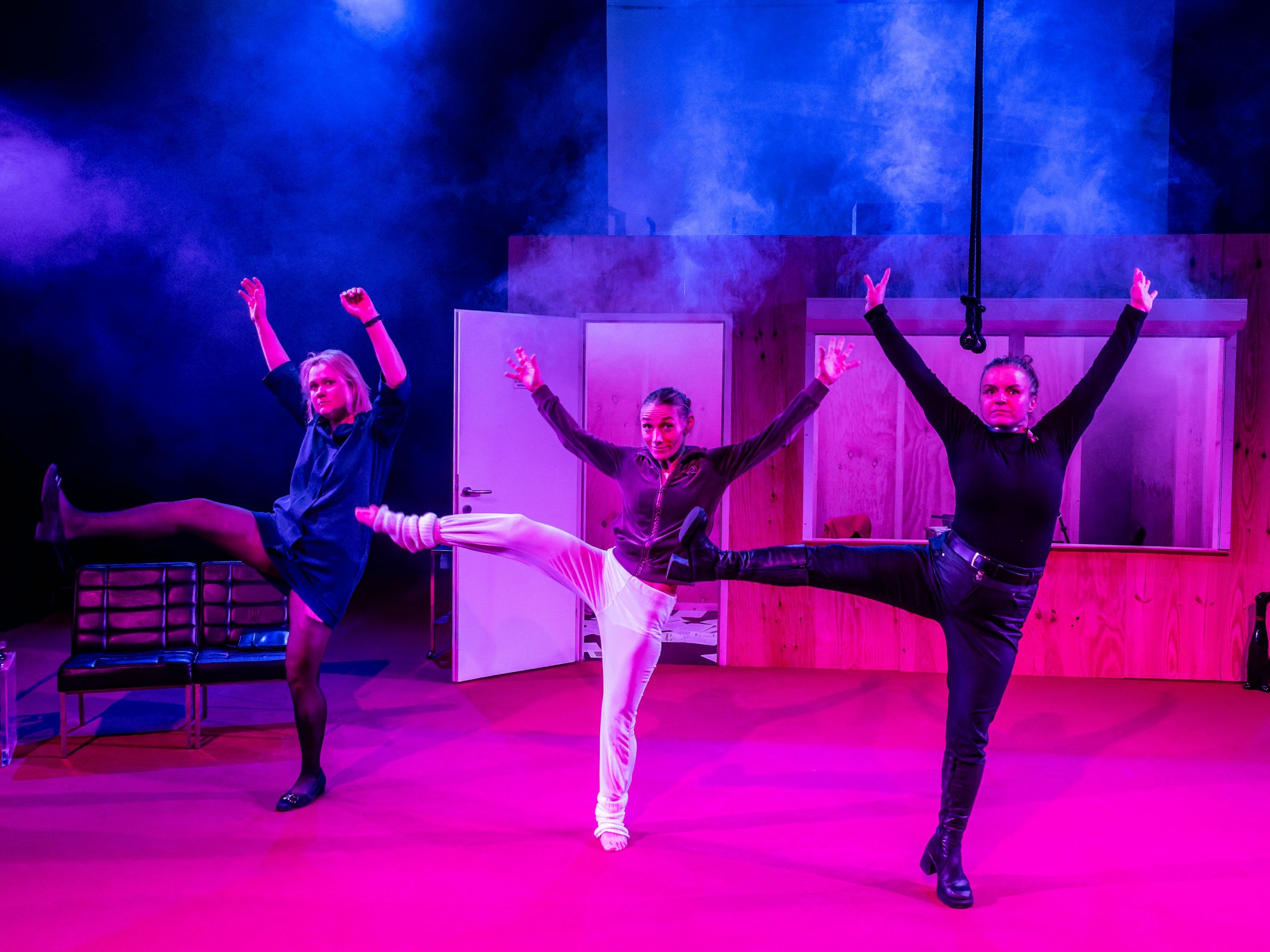 Three performers striking dynamic poses on stage, surrounded by purple lighting and smoke, with a set featuring wooden structures and benches.