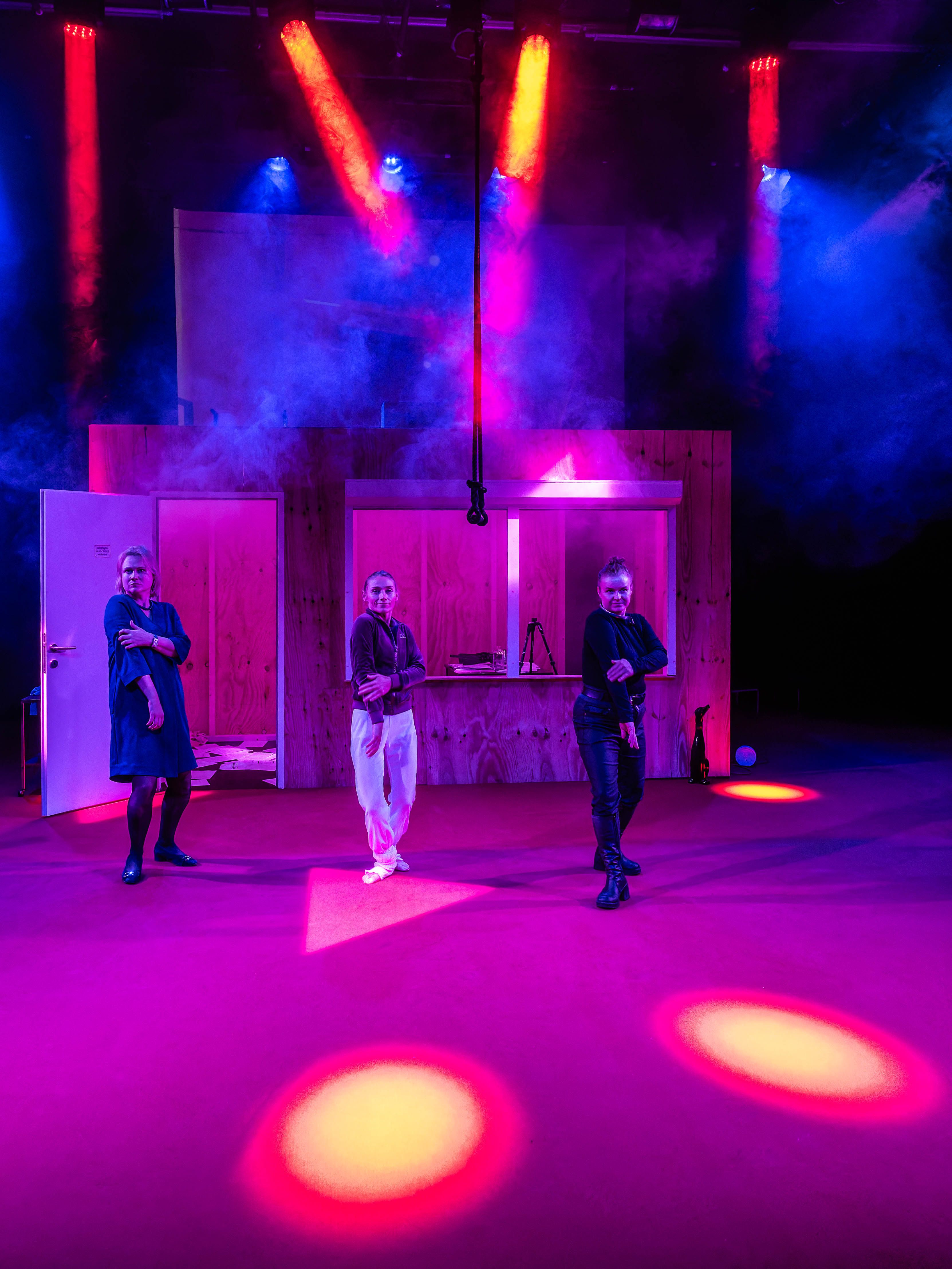 Three performers in stylish outfits pose on a brightly lit stage, surrounded by colourful lighting and smoke effects, with wooden set pieces in the background.