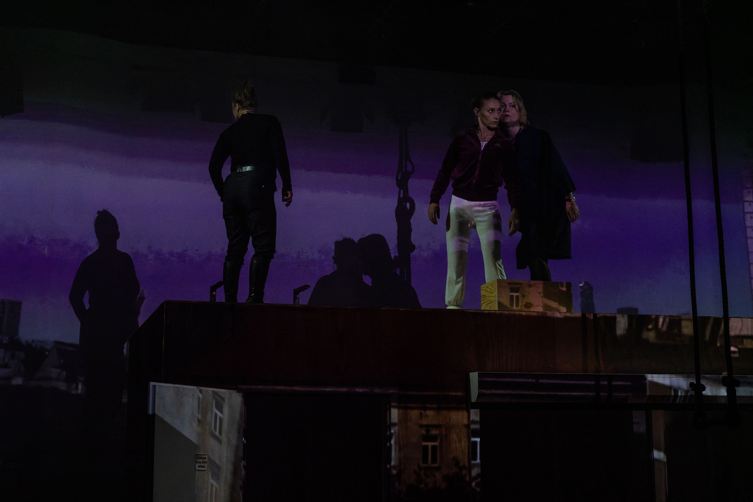 Two performers on a stage with a purple projected background, one standing and one leaning close together, with shadows visible in the scene