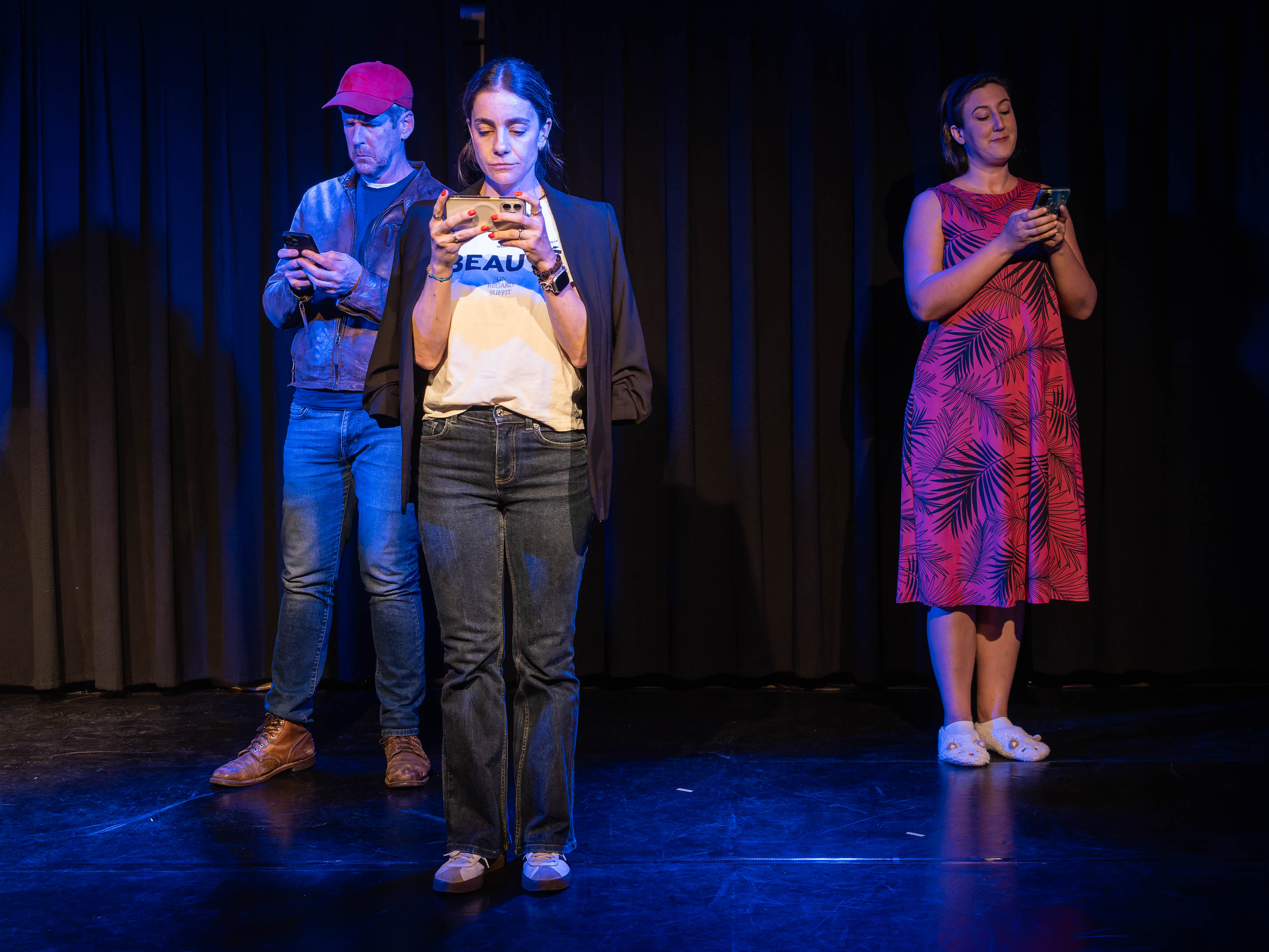 Three performers standing on stage, each focused on their mobile phones. The first person, wearing a red cap and a jacket, is on the left. The second person in a white t-shirt and black blazer stands in the middle. The third person, in a pink dress with a leaf pattern, stands on the right. The background features black curtains and blue stage lighting.