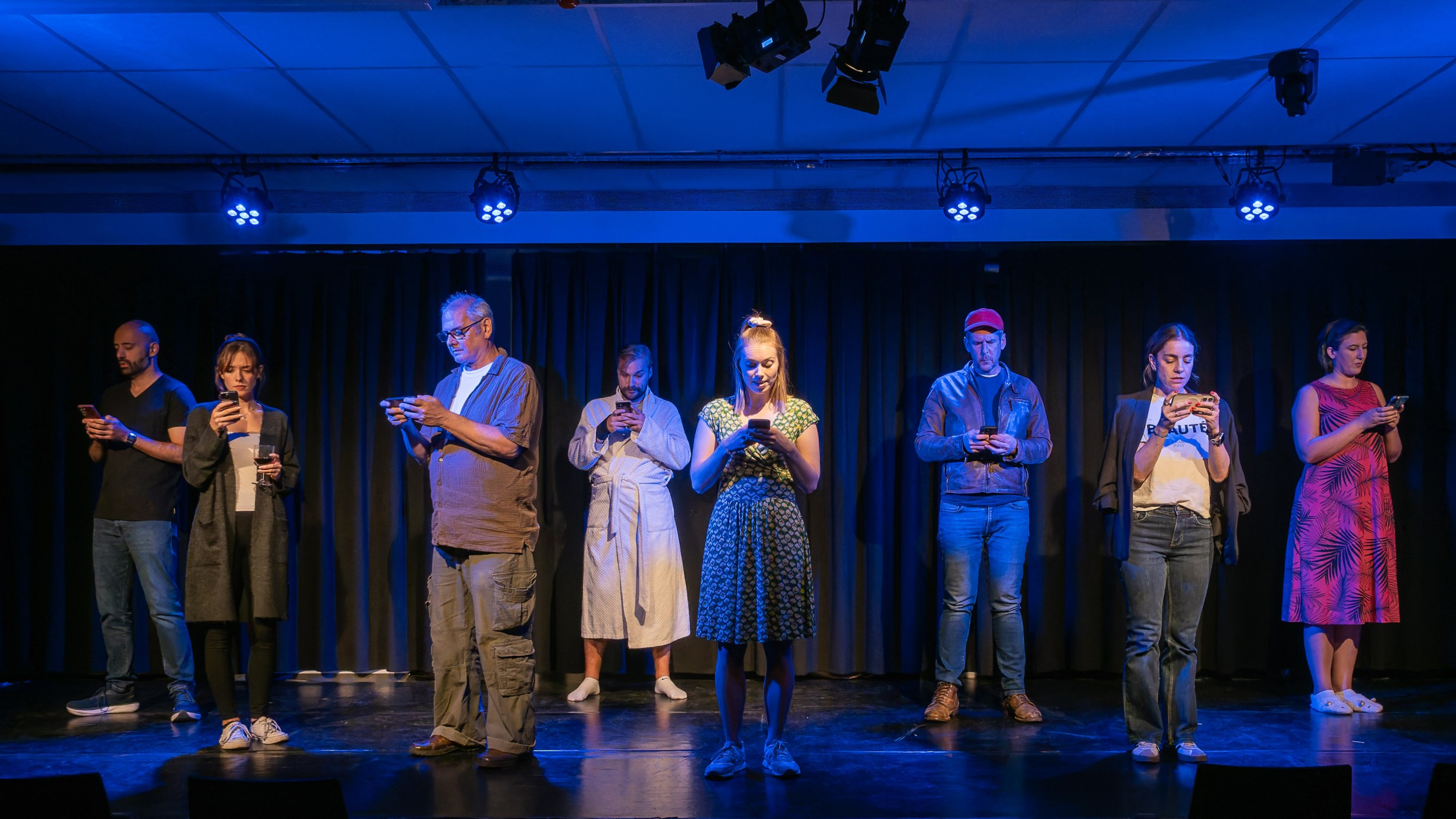 A group of nine people stand on stage, each focused on their smartphones, with blue stage lighting and a black curtain in the background.