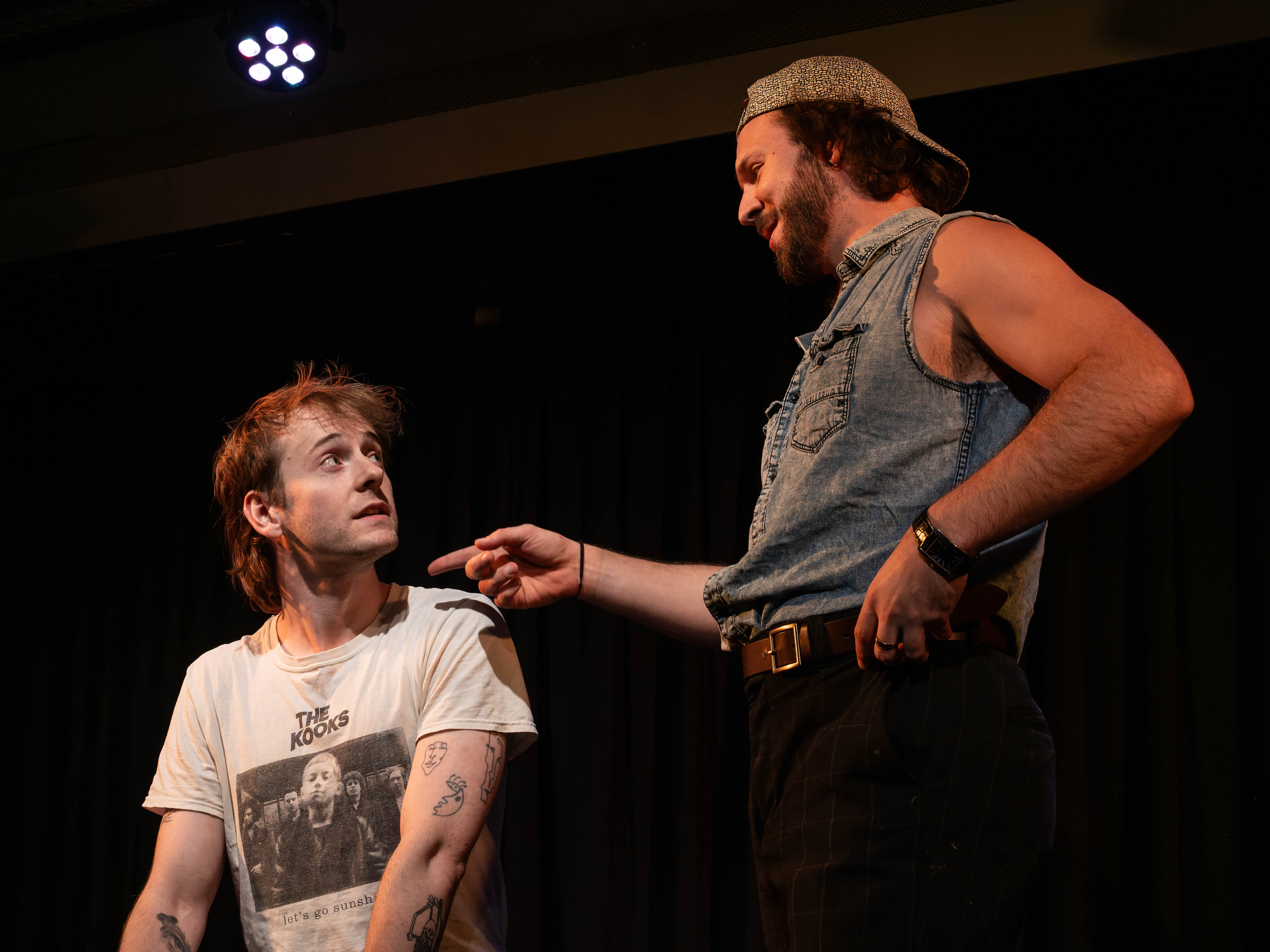 Two male performers on stage in a theatrical setting. One, seated and wearing a T-shirt with a band print, looks up with a surprised expression, while the other, standing and dressed in a sleeveless denim top, points playfully at him.