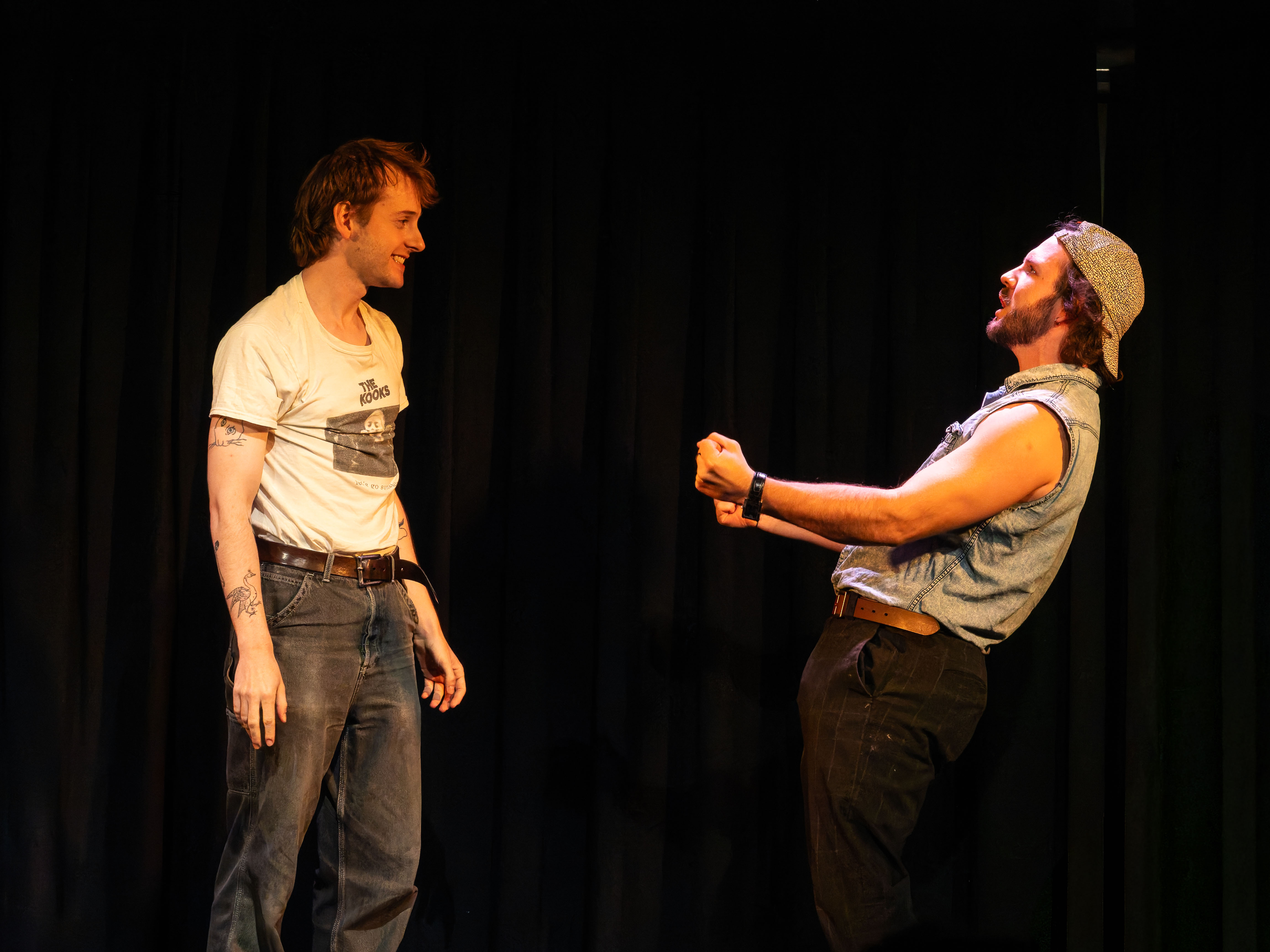 Two male performers on stage, one smiling and the other expressing a dramatic pose, against a black curtain backdrop.