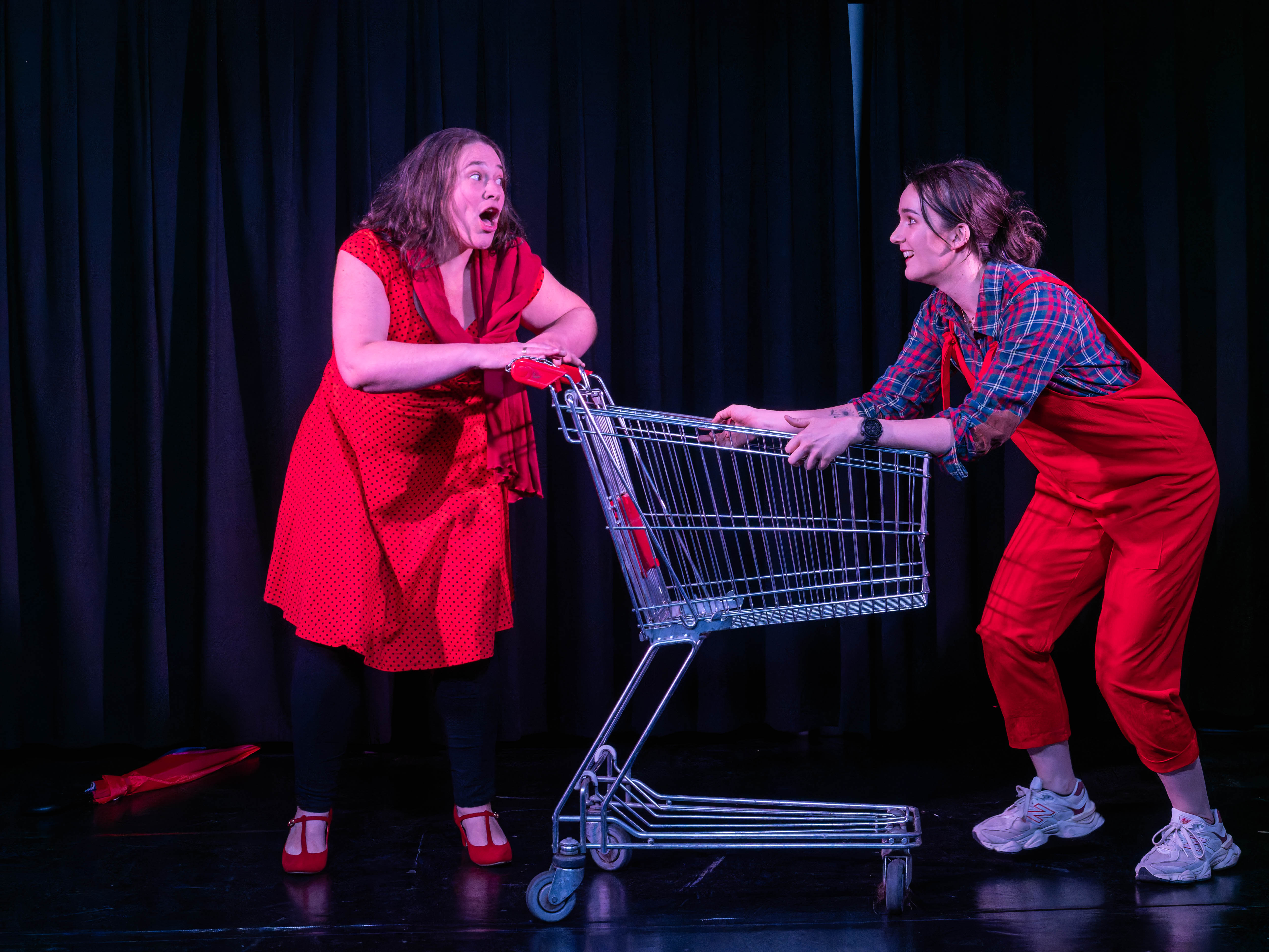 Two women performing on stage, one in a red polka dot dress and the other in red overalls, interacting playfully with a shopping trolley against a black backdrop.
