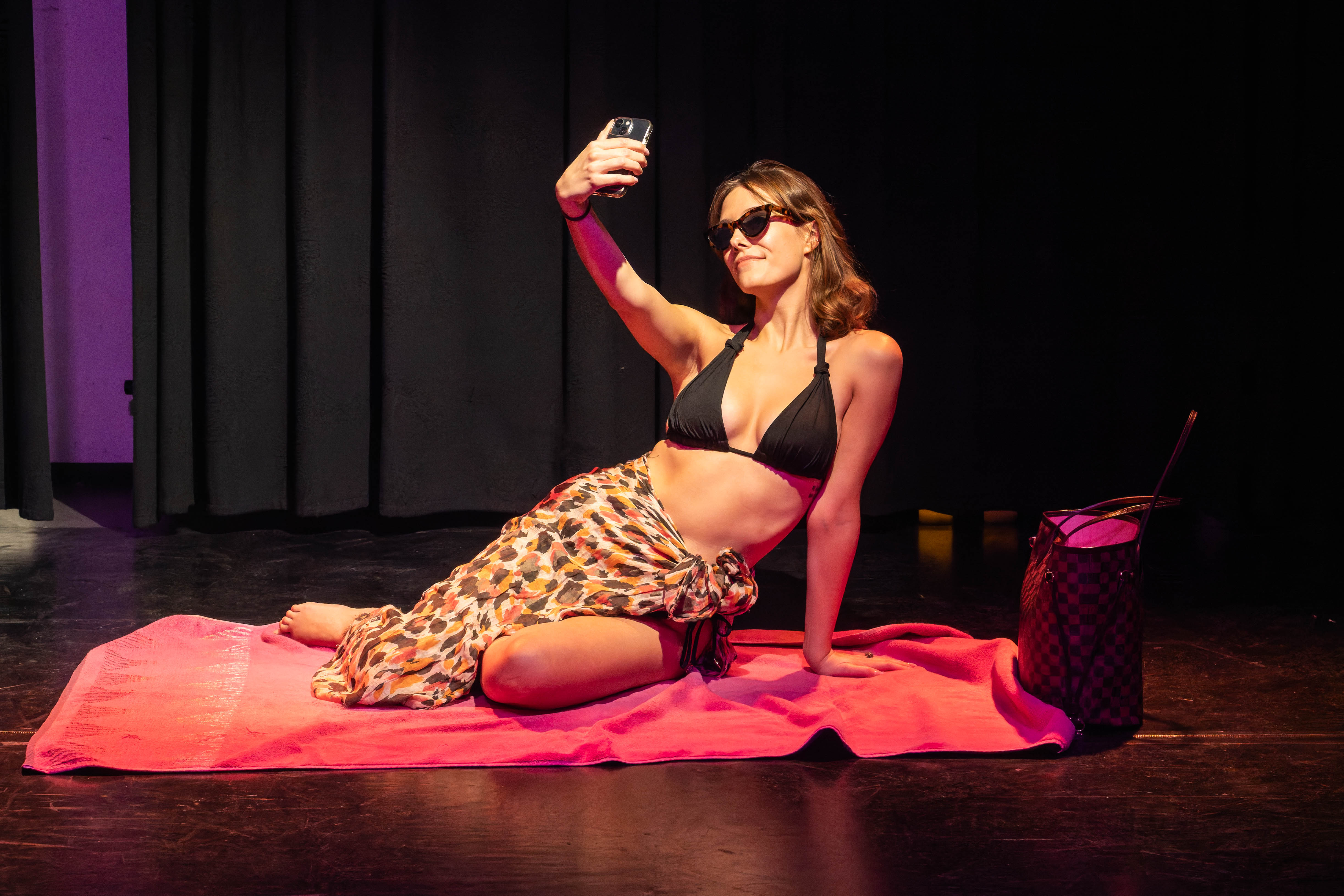 A woman in a black bikini and sunglasses sits on a pink towel, taking a selfie with her phone. She has a colourful sarong wrapped around her waist and a stylish bag beside her. The background features dark curtains.