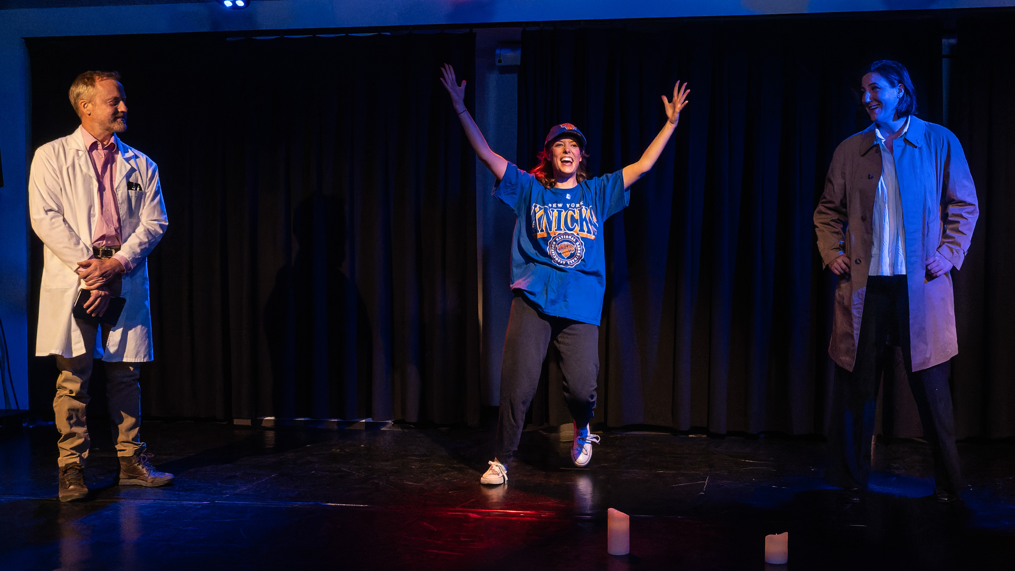 A scene from a theatre production featuring a woman in a blue New York Knicks t-shirt and cap performing energetically in the centre, while a male actor in a lab coat and another in a long coat observe her with smiles.