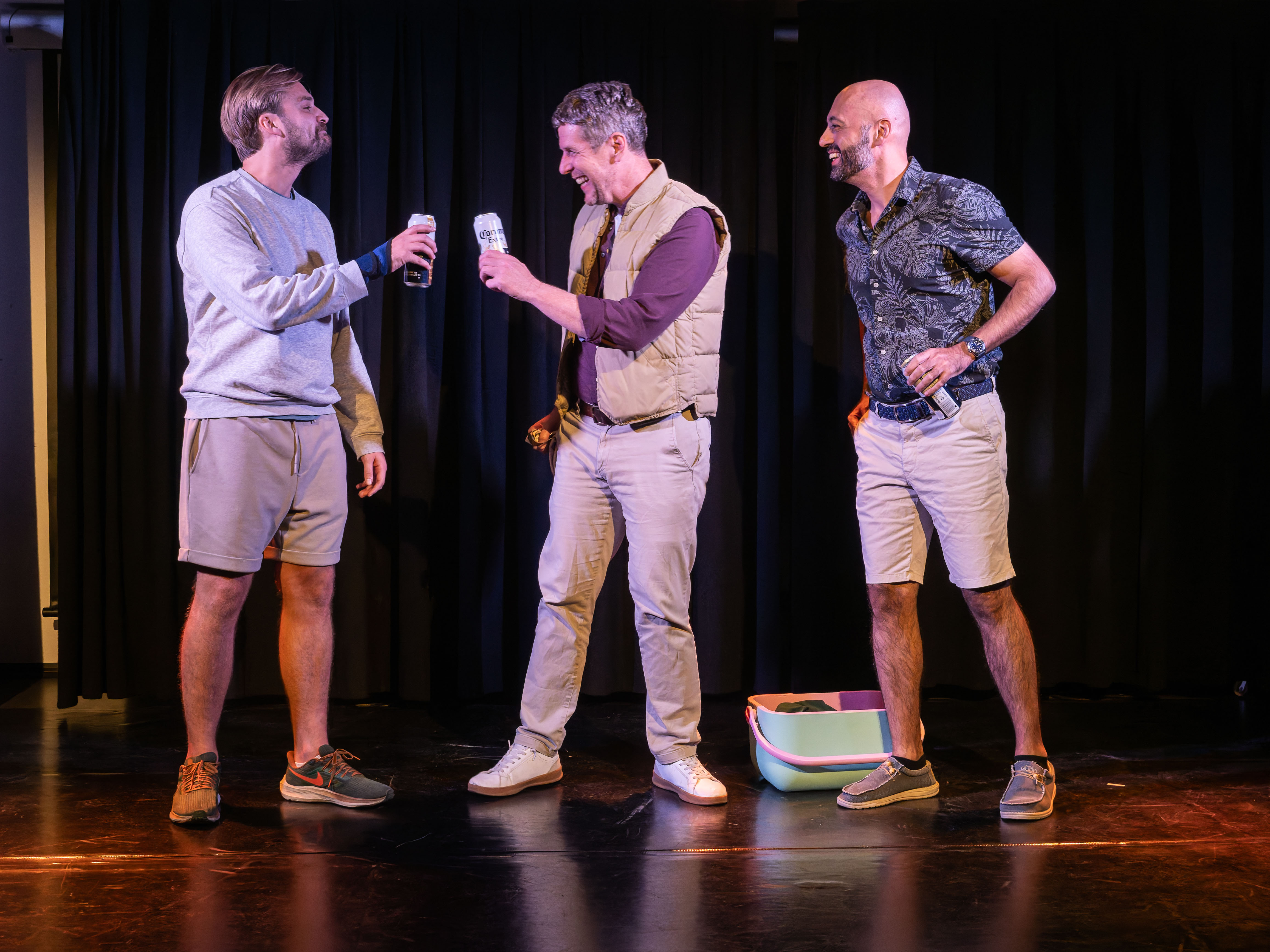 Three men engaged in a toast on stage, holding cans in their hands. They are smiling and dressed casually in a performance setting with a dark curtain backdrop.