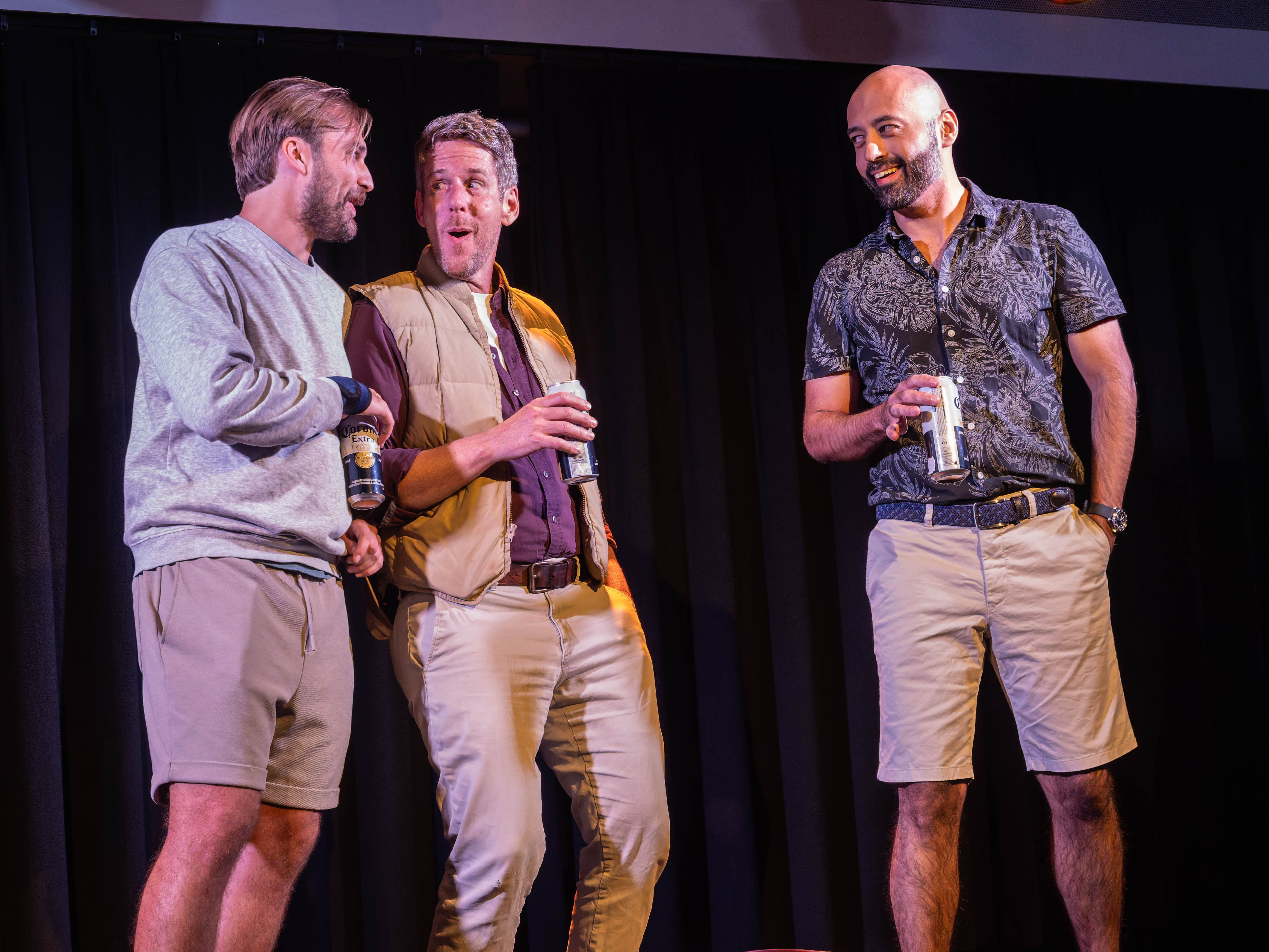 Three men on stage, two holding cans of beer and one holding a drink, engaged in conversation and smiling, against a black curtain backdrop.