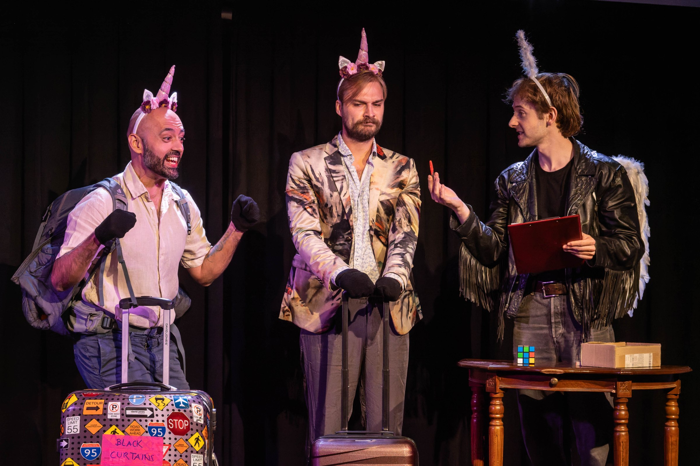 Three performers on stage, two wearing unicorn headbands and one with angel wings, engaged in a comedic scene. One is animatedly expressing excitement next to a suitcase covered in stickers, while another stands with a serious expression, and the third holds a clipboard.