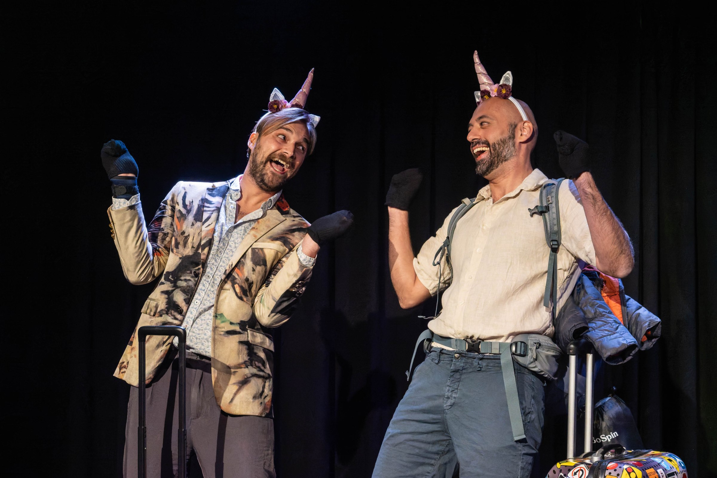 Two men in playful poses, wearing unicorn headbands, with one dressed in a patterned blazer and the other in a light shirt. Both are smiling and wearing black gloves, with a suitcase and a backpack beside them.