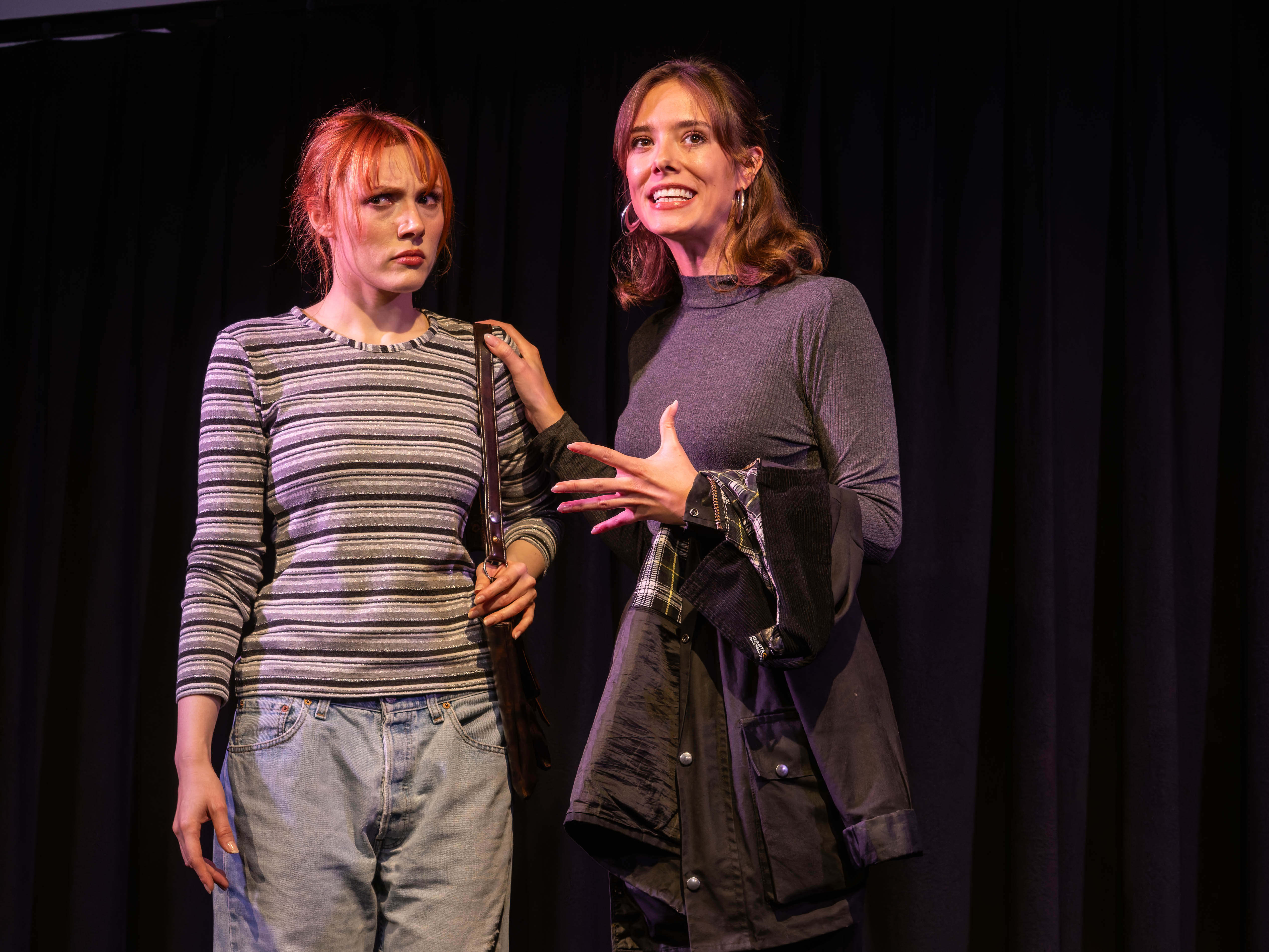 Two women in casual clothing are performing on stage, with one woman looking concerned and the other animatedly expressing herself, against a black curtain backdrop.