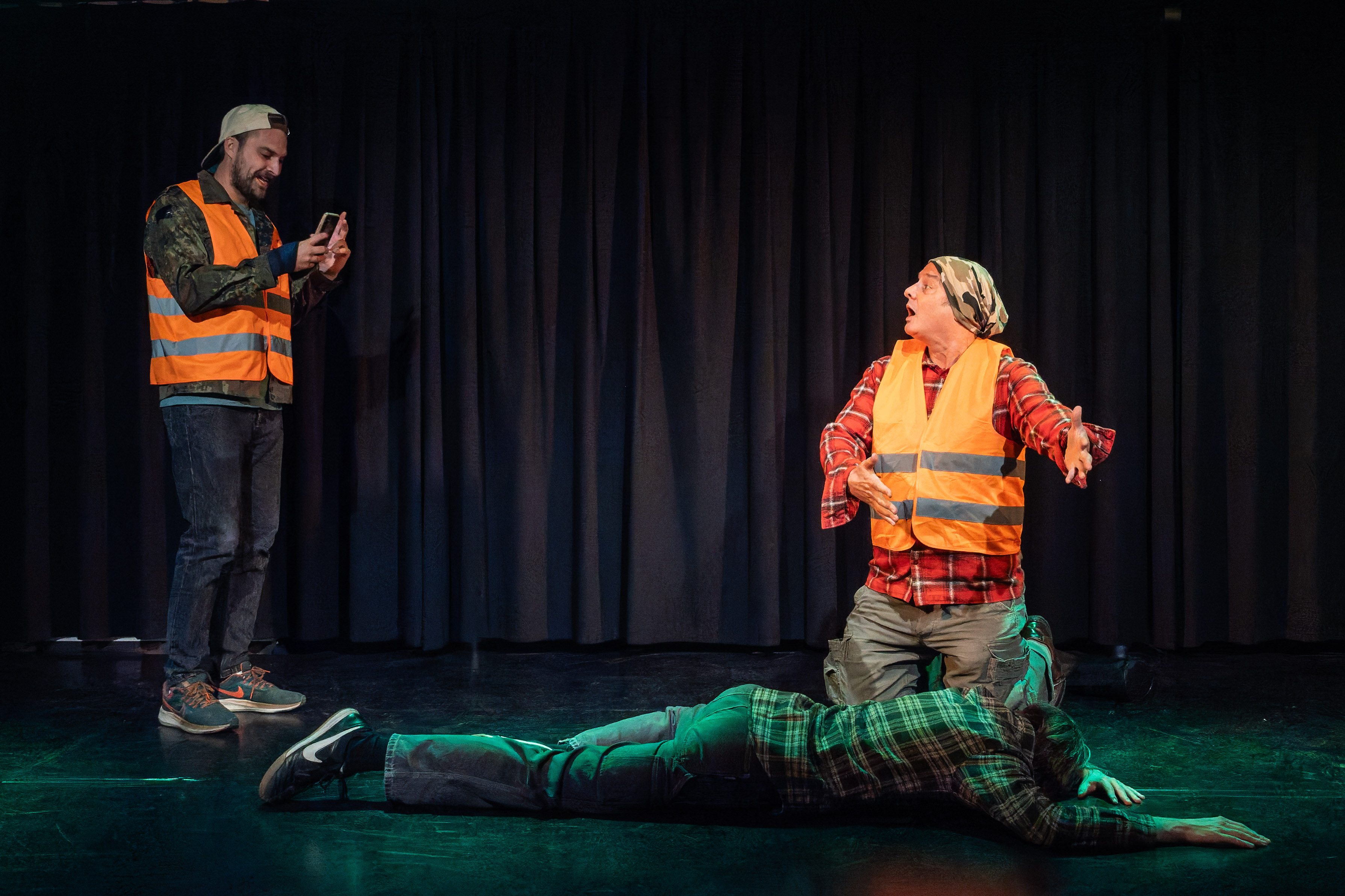 A scene from a theatrical performance featuring two actors in high-visibility vests, one standing and using a smartphone, while the other kneels beside a person lying on the ground.