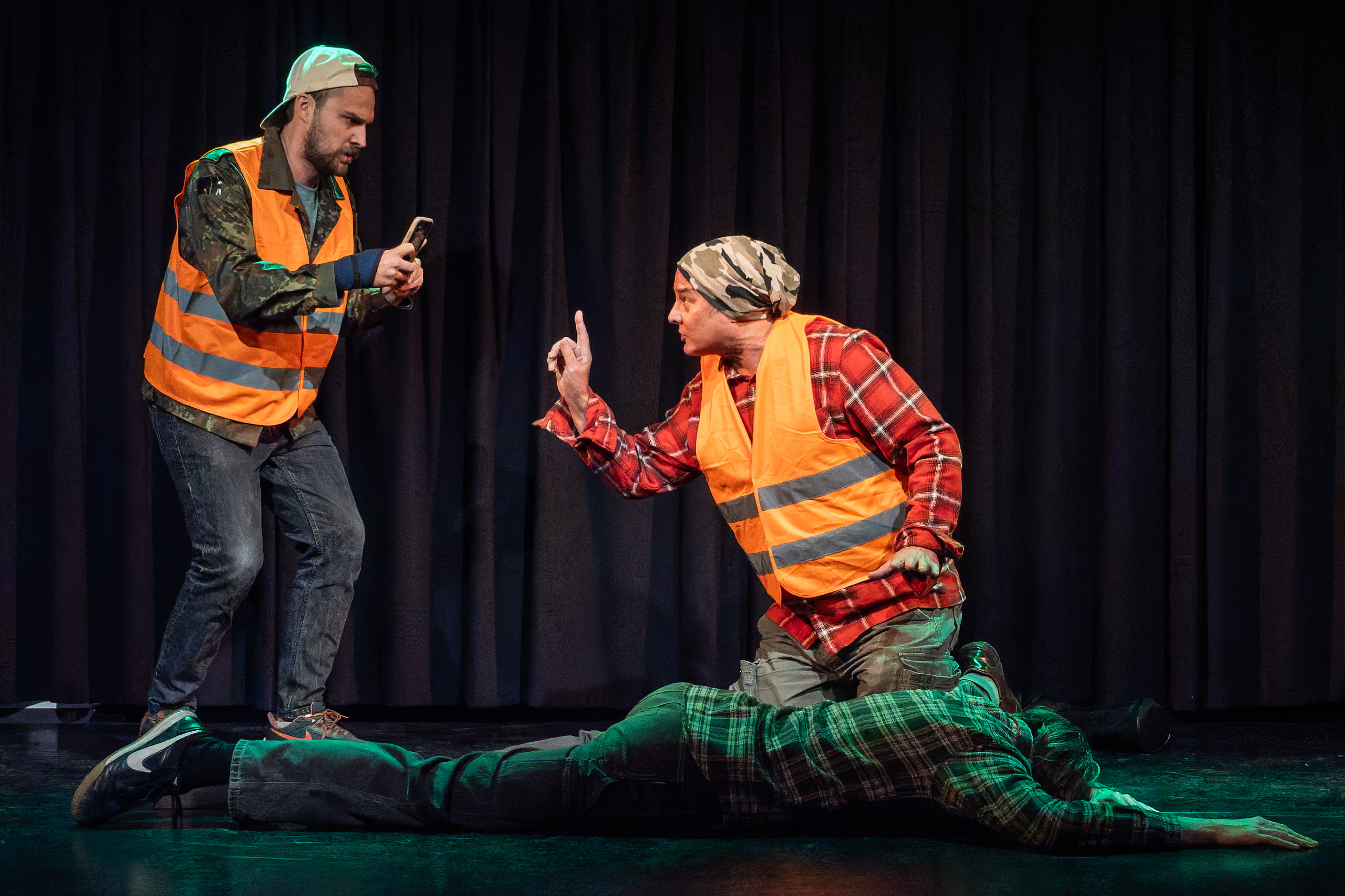 Two actors in high-visibility vests perform on stage, one taking a photo with a mobile phone, while the other gestures emphatically. A third actor lies on the ground in a plaid shirt.