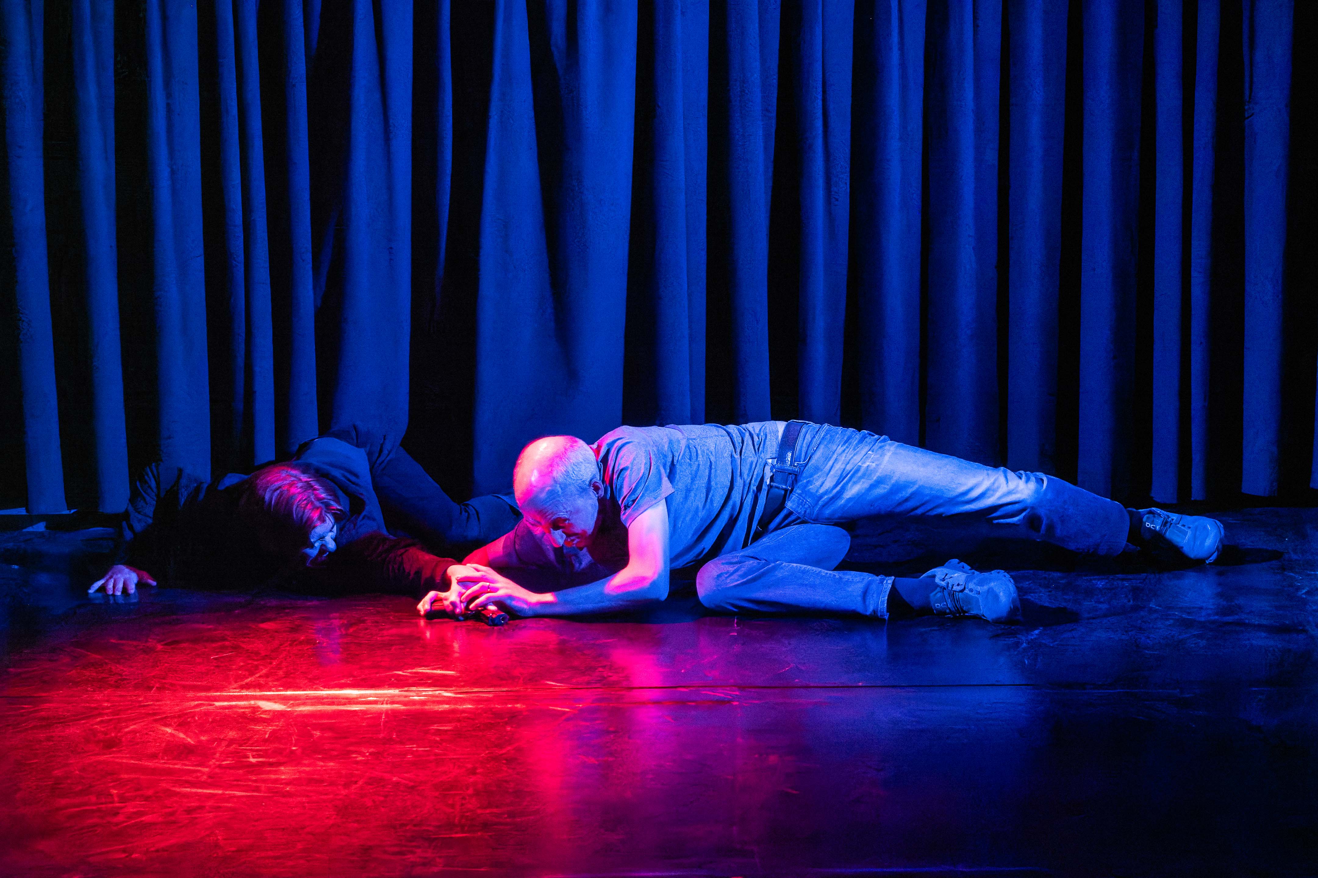 A dramatic scene featuring two performers on a dimly lit stage, one lying on the ground reaching out, while the other is crawling towards them. The background is a curtain, and the lighting creates a blue and pink hue.