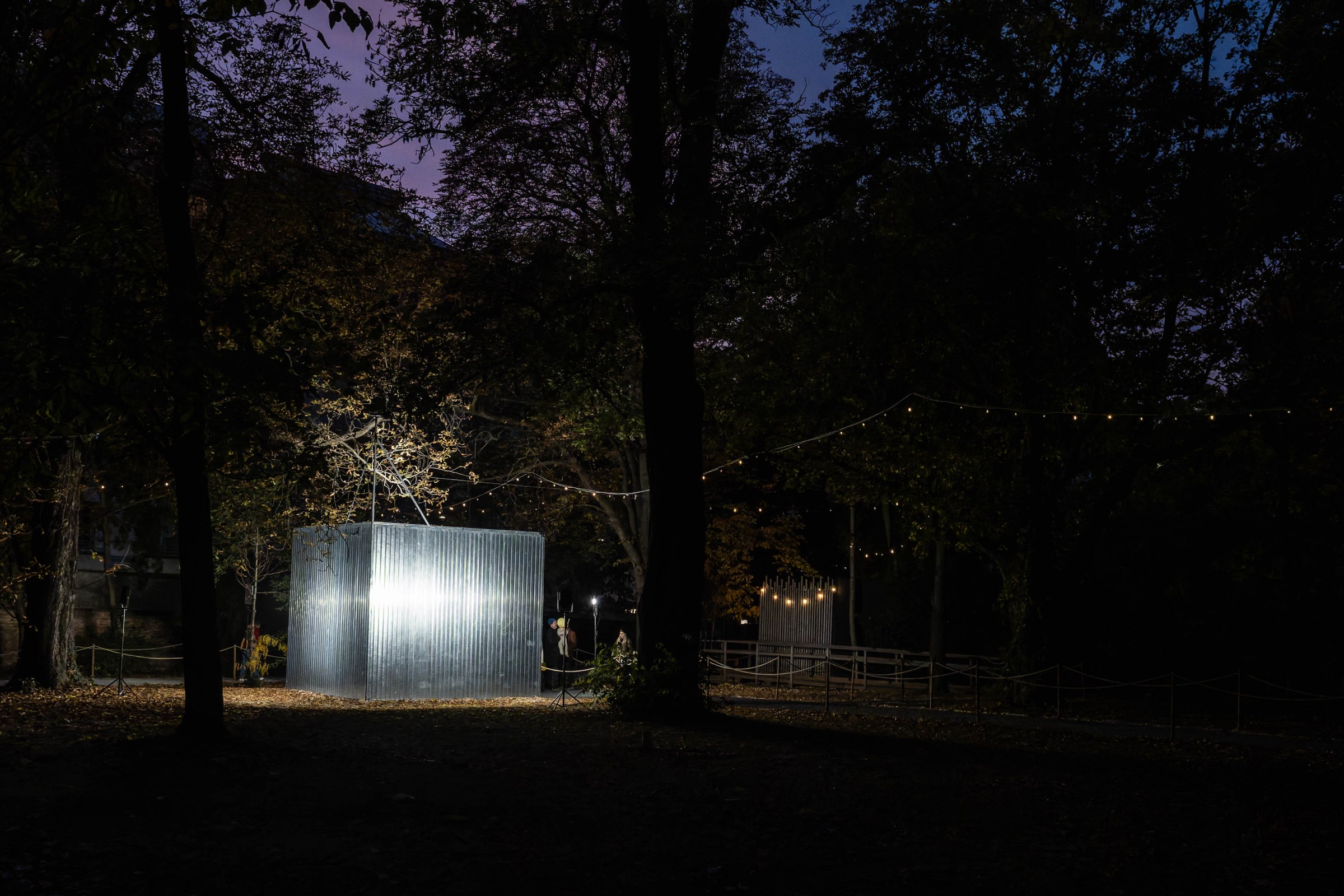 A lit metallic structure standing in a dark park surrounded by trees, with warm string lights illuminating the area.