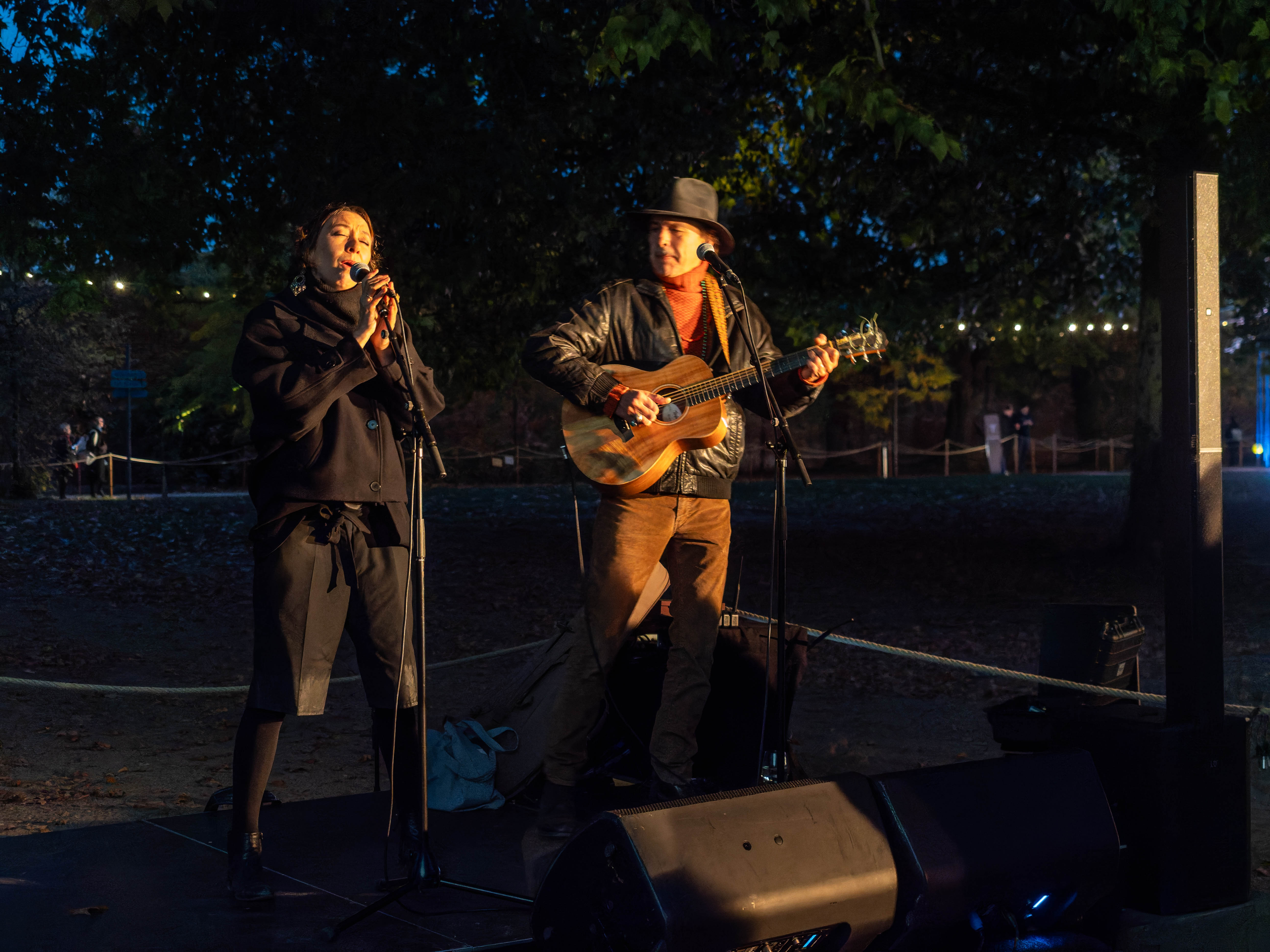 A woman singing into a microphone while a man plays guitar beside her, both illuminated by soft lighting in a park setting during dusk.