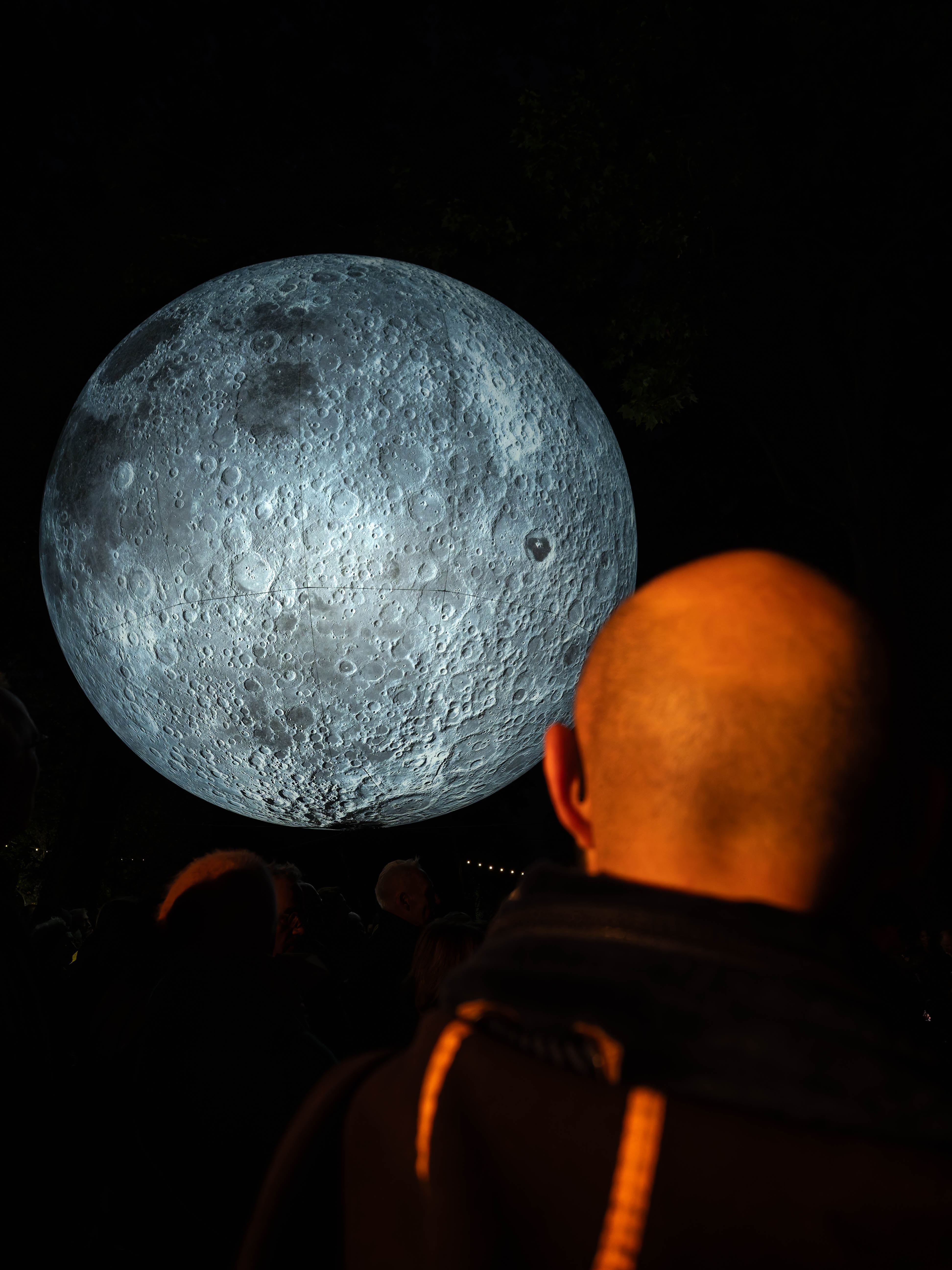 A large illuminated model of the Moon hangs in a dark setting, with a crowd of people visible in the foreground, observing its detailed surface features.