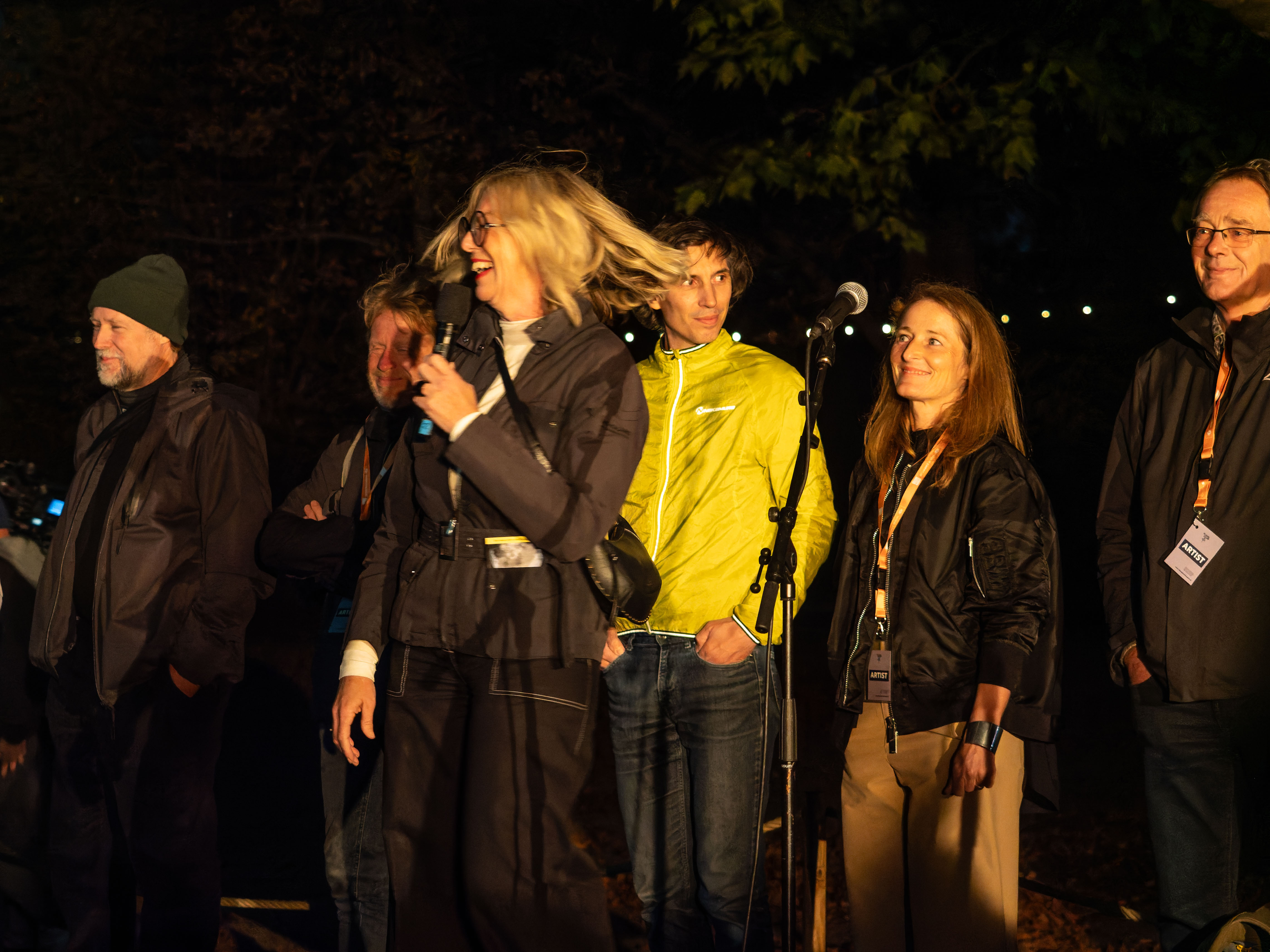 A group of five people standing on stage during an evening event, with one woman speaking into a microphone. The setting is outdoors with soft lighting and trees in the background.