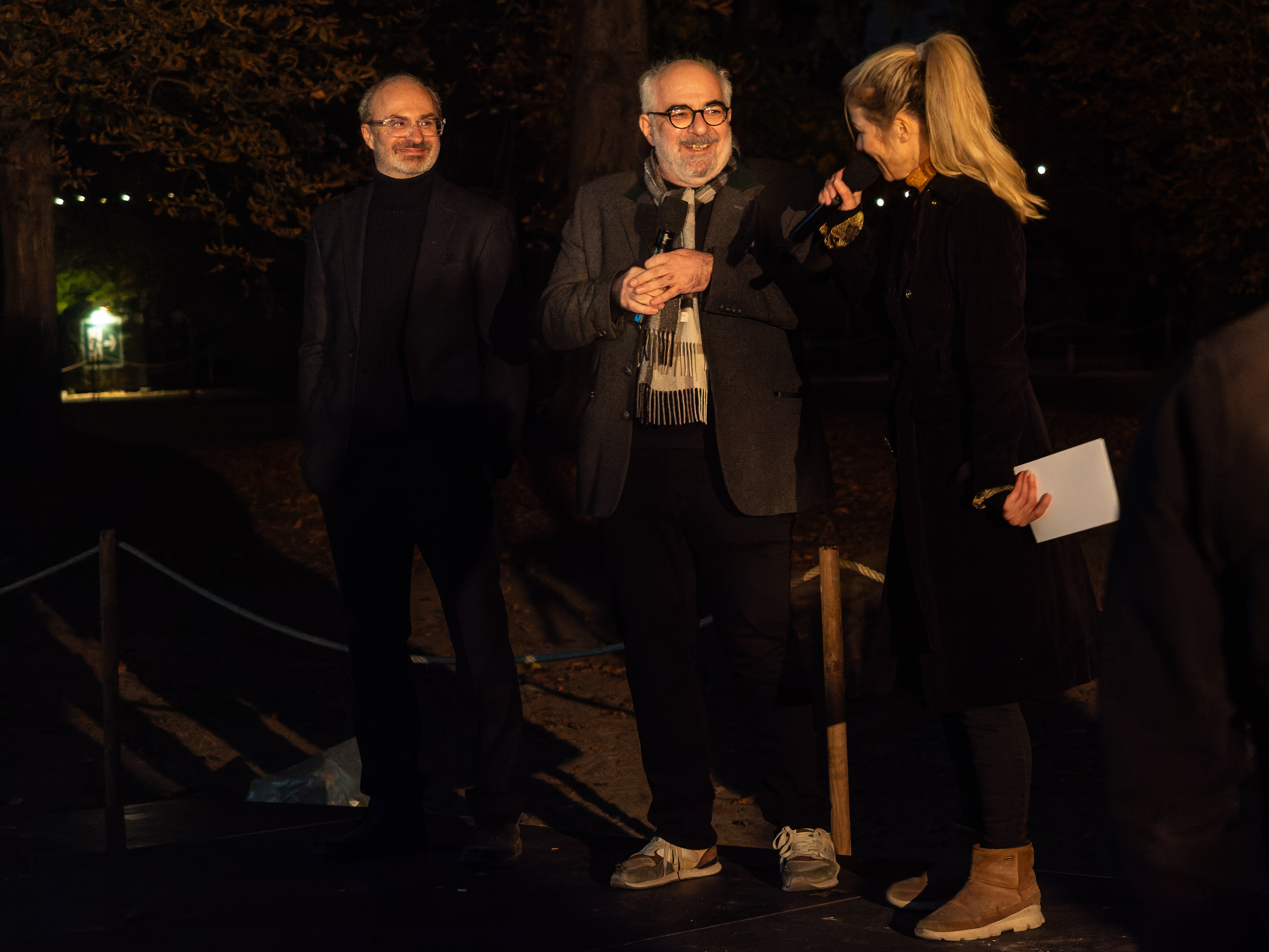 Three individuals conversing on a stage at night, with trees in the background and soft lighting in the distance.