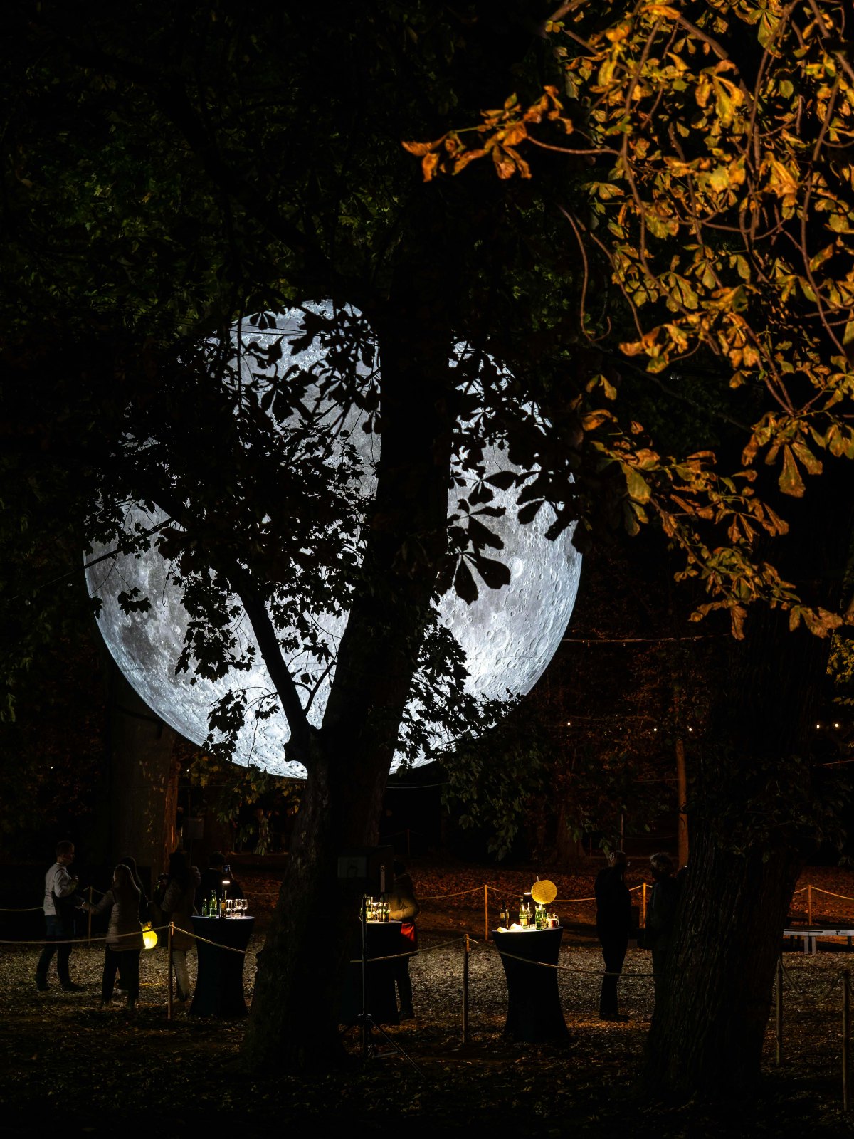 A large illuminated moon installation is displayed in a wooded area at night, with guests mingling around tables adorned with drinks.
