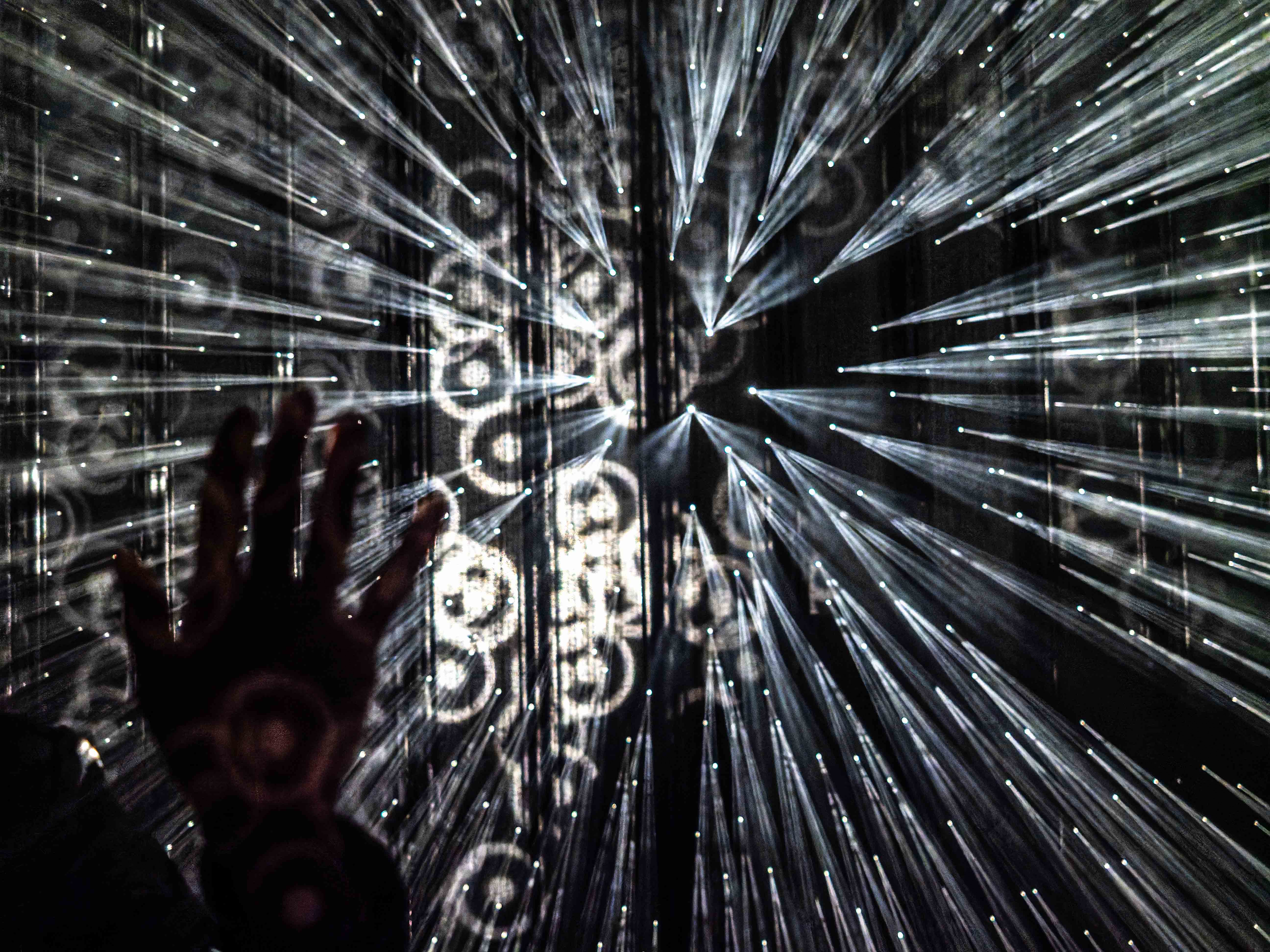 A hand reaching towards a projection of light beams and circular patterns, creating a dynamic visual effect.