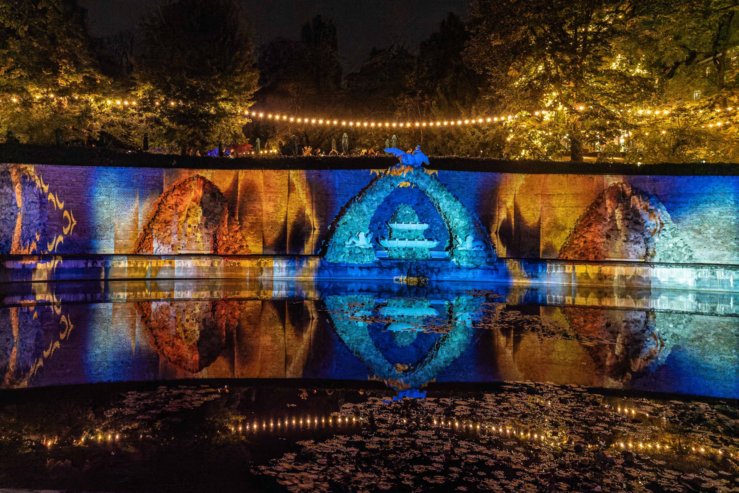 A beautifully illuminated garden at night, featuring a water feature with reflections, surrounded by multi-coloured lighting on a stone wall, and string lights overhead.