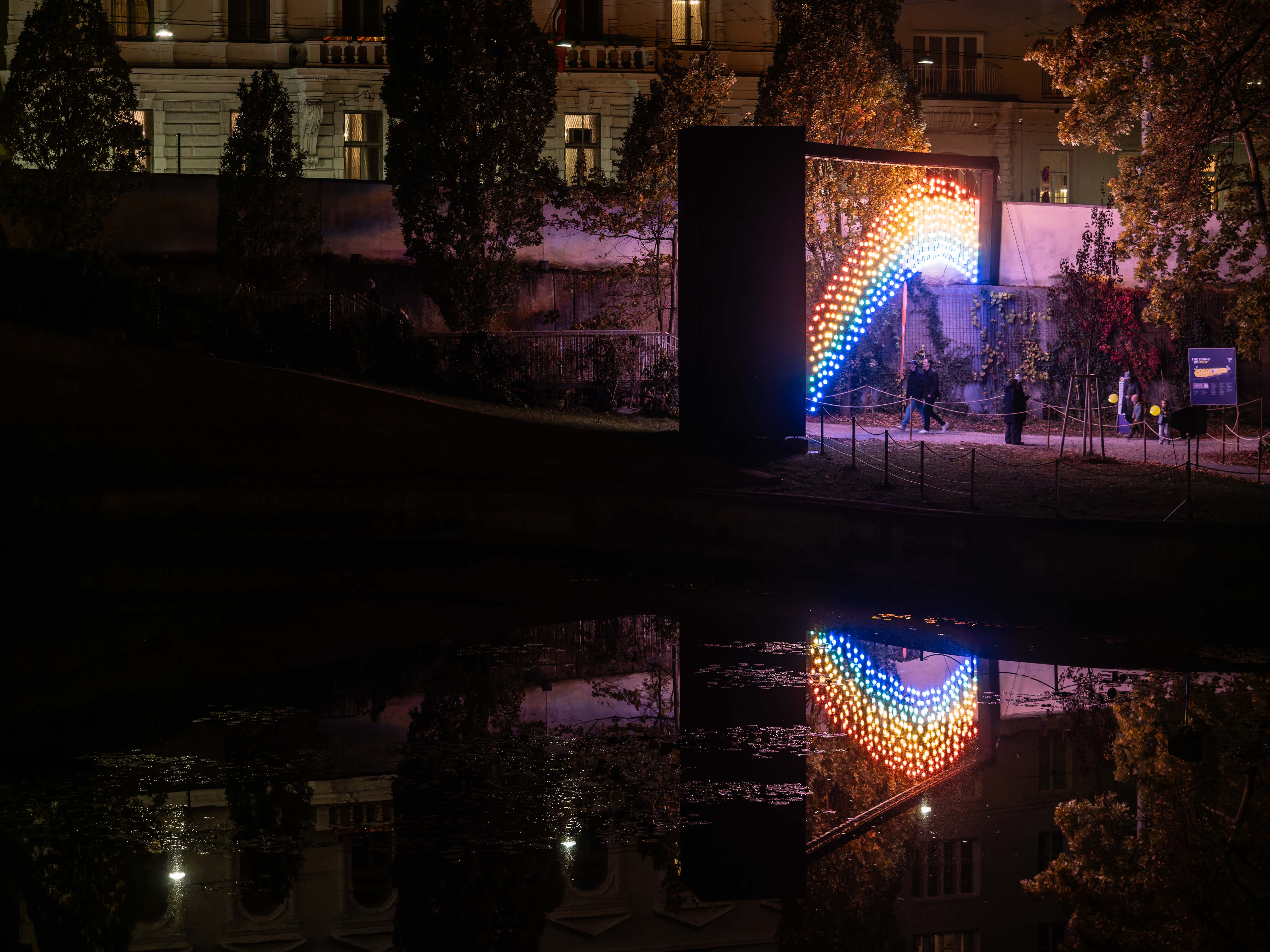 An illuminated archway featuring colourful lights reflecting in a calm body of water at night, with people walking nearby.