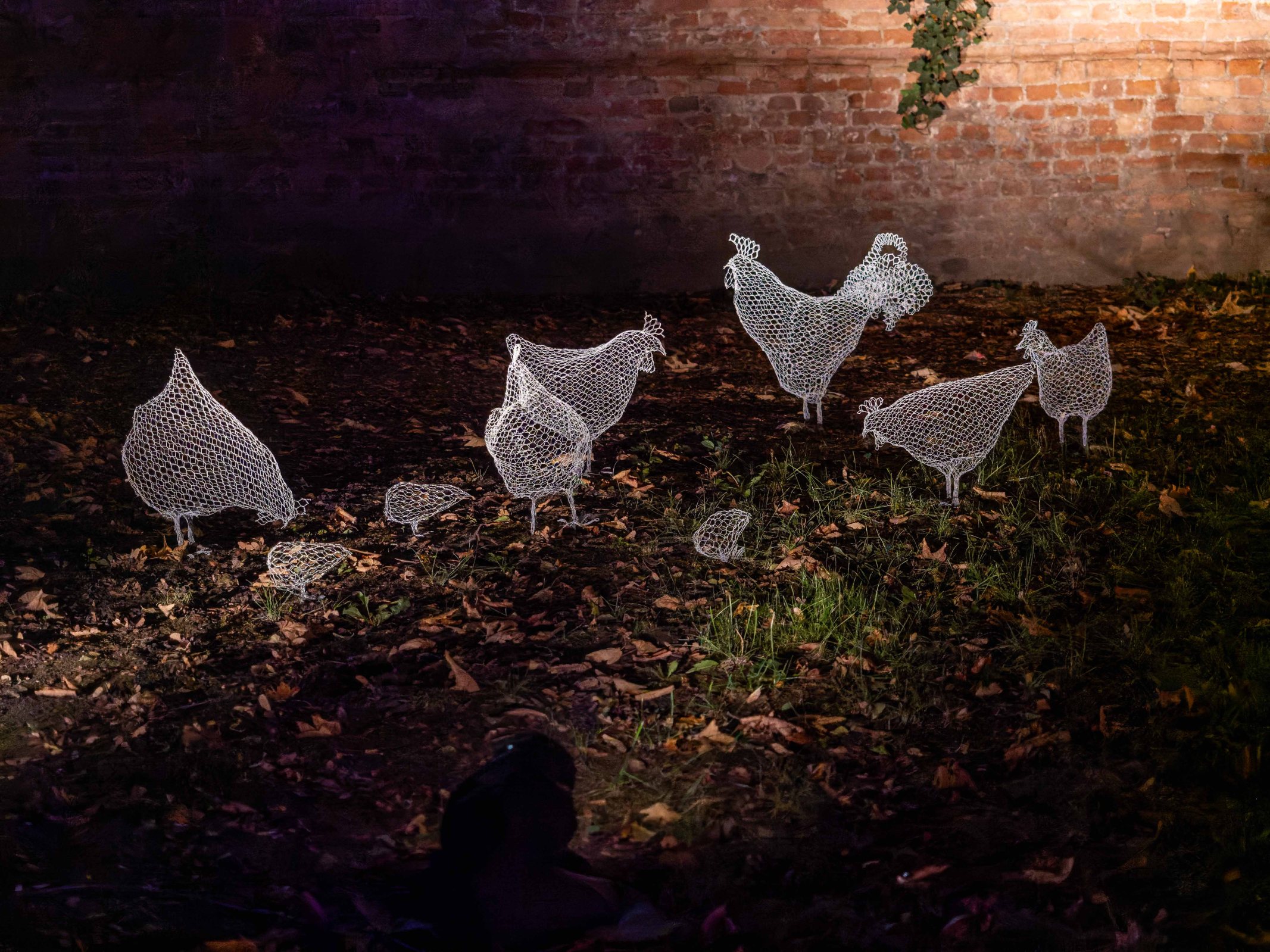 A series of wireframe chicken sculptures arranged on the ground amidst fallen leaves, set against a brick wall.