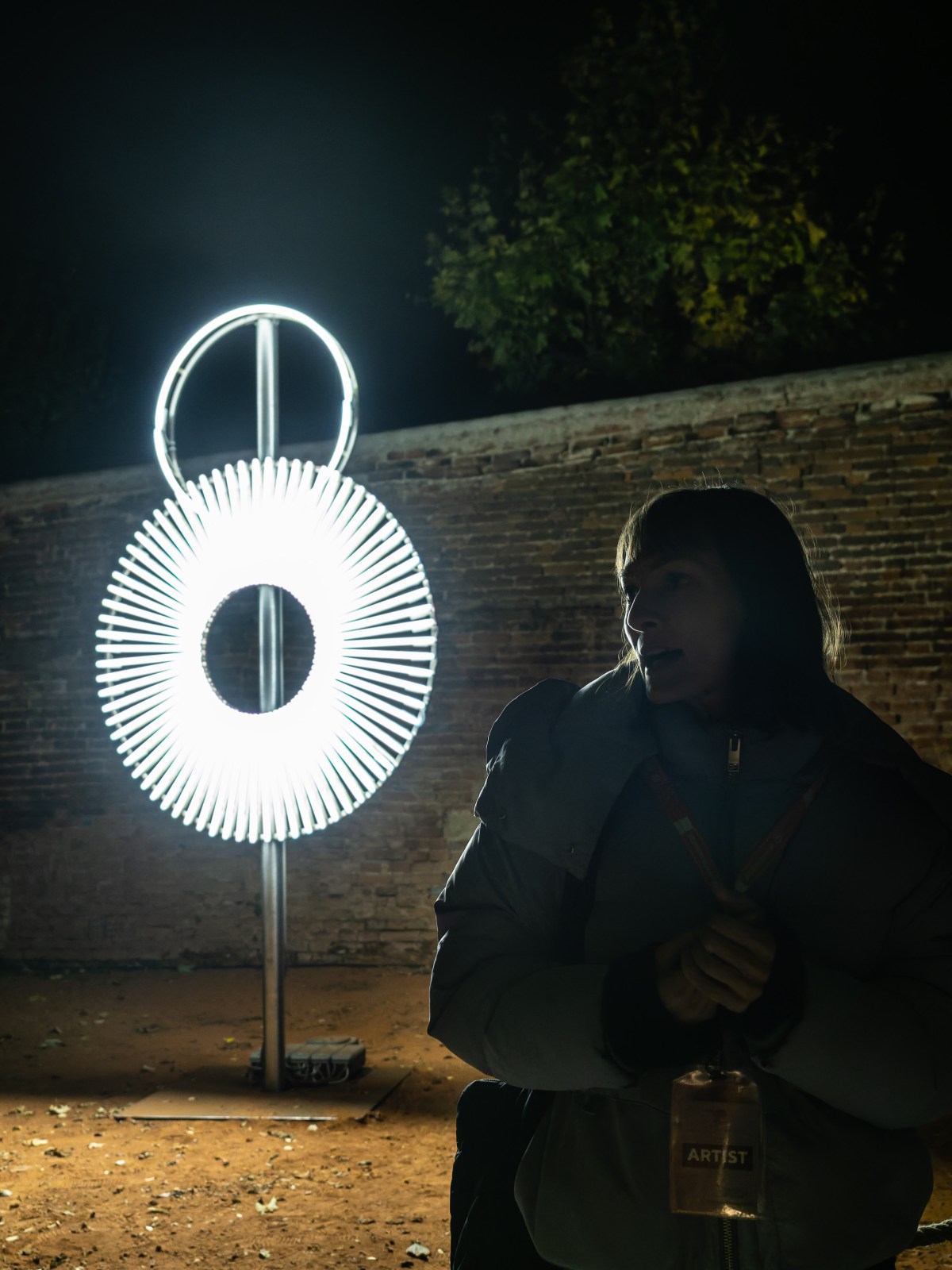 A woman in a light grey coat stands in front of a circular light installation at night, with illuminated circular patterns contrasting against a dark background.