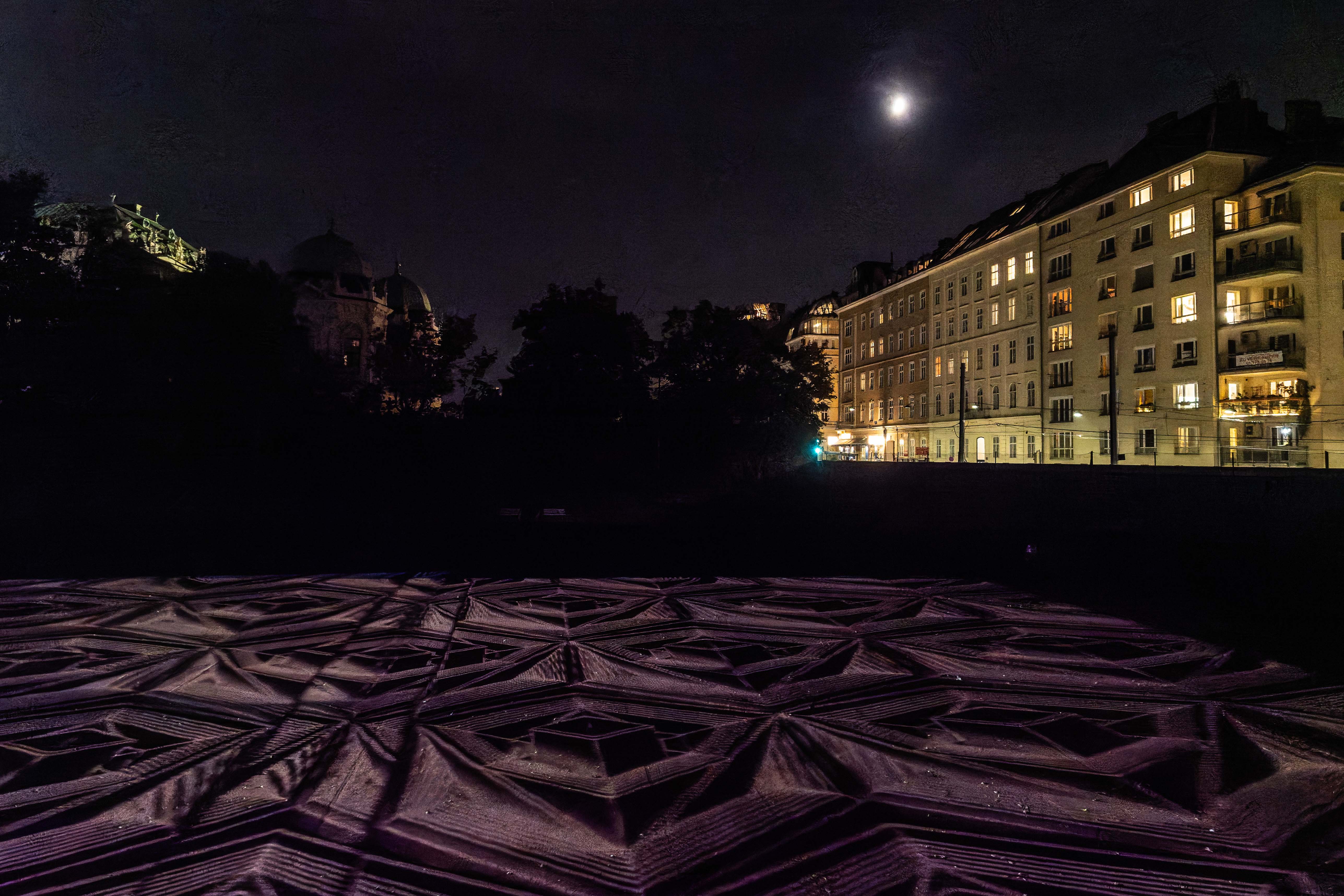 Night scene featuring a patterned surface illuminated by moonlight, with a backdrop of low-rise buildings on one side and trees on the other.