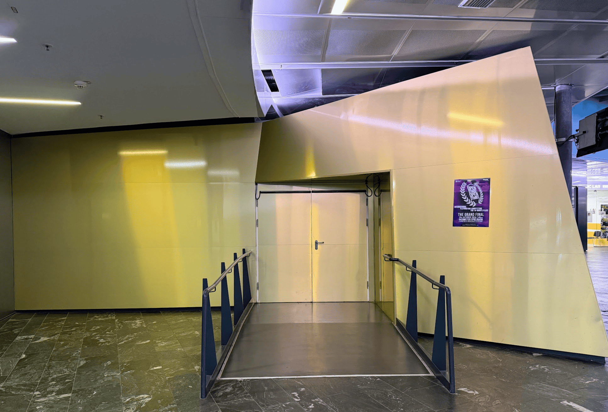 A modern, angled entrance door with a ramp and handrails, featuring glossy yellow walls and a marble floor.